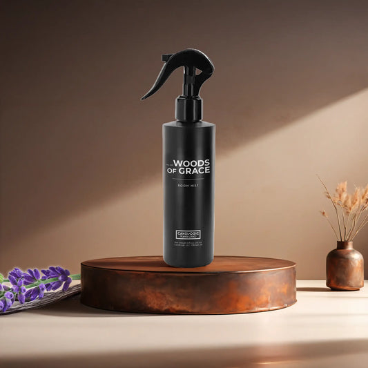 Candl•ogic’s 8 oz. room mist in a black spray bottle labeled ‘No. 001 Woods of Grace’ on a copper tray with lavender and vanilla beans and a copper vase in the background.