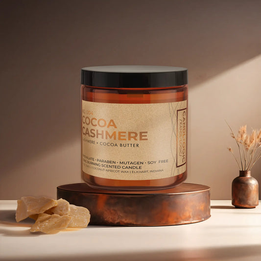 Candl•ogic’s scented candle with a wood wick in an amber glass 16 oz. jar labeled ‘No. 004 Cocoa Cashmere’ on a copper tray with an chunks of cocoa butter and a copper vase in the background.