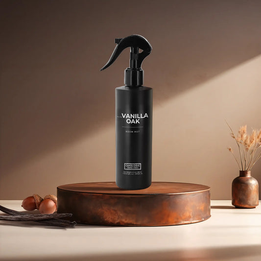 Candl•ogic’s 8 oz. room mist in a black spray bottle labeled ‘No. 034 Vanilla Oak’ on a copper tray with acorns and vanilla beans and a copper vase in the background.