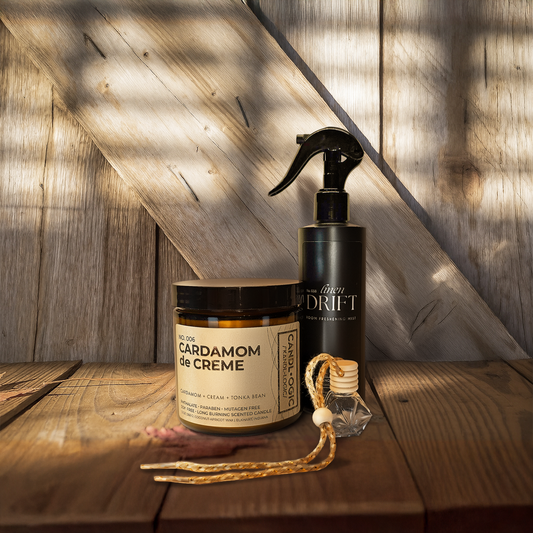 Home fragrance products including a scented candle, room spray bootle and a car diffuser on a wooden surface with a wooden wall background.