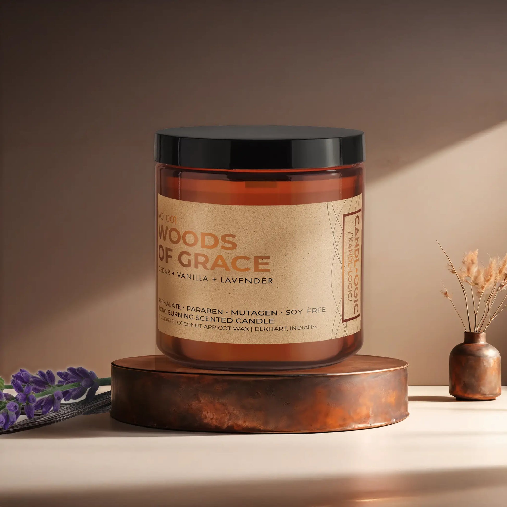 Candl•ogic’s scented candle with a wood wick in an amber glass 16 oz. jar labeled ‘No. 001 Woods of Grace’ on a copper tray with lavender and vanilla beans and a copper vase in the background.
