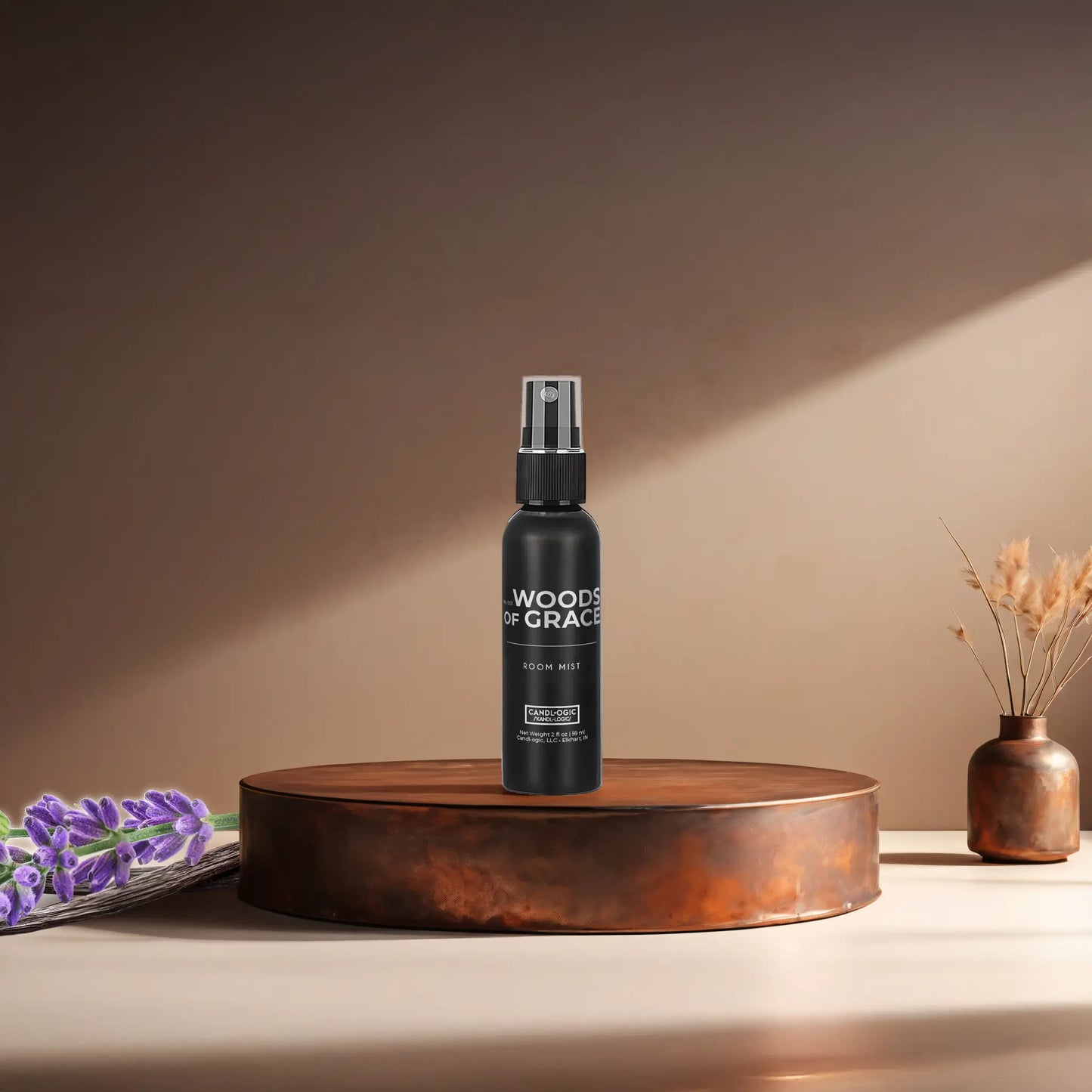 Candl•ogic’s 2 oz. room mist in a black spray bottle labeled ‘No. 001 Woods of Grace’ on a copper tray with lavender and vanilla beans and a copper vase in the background.