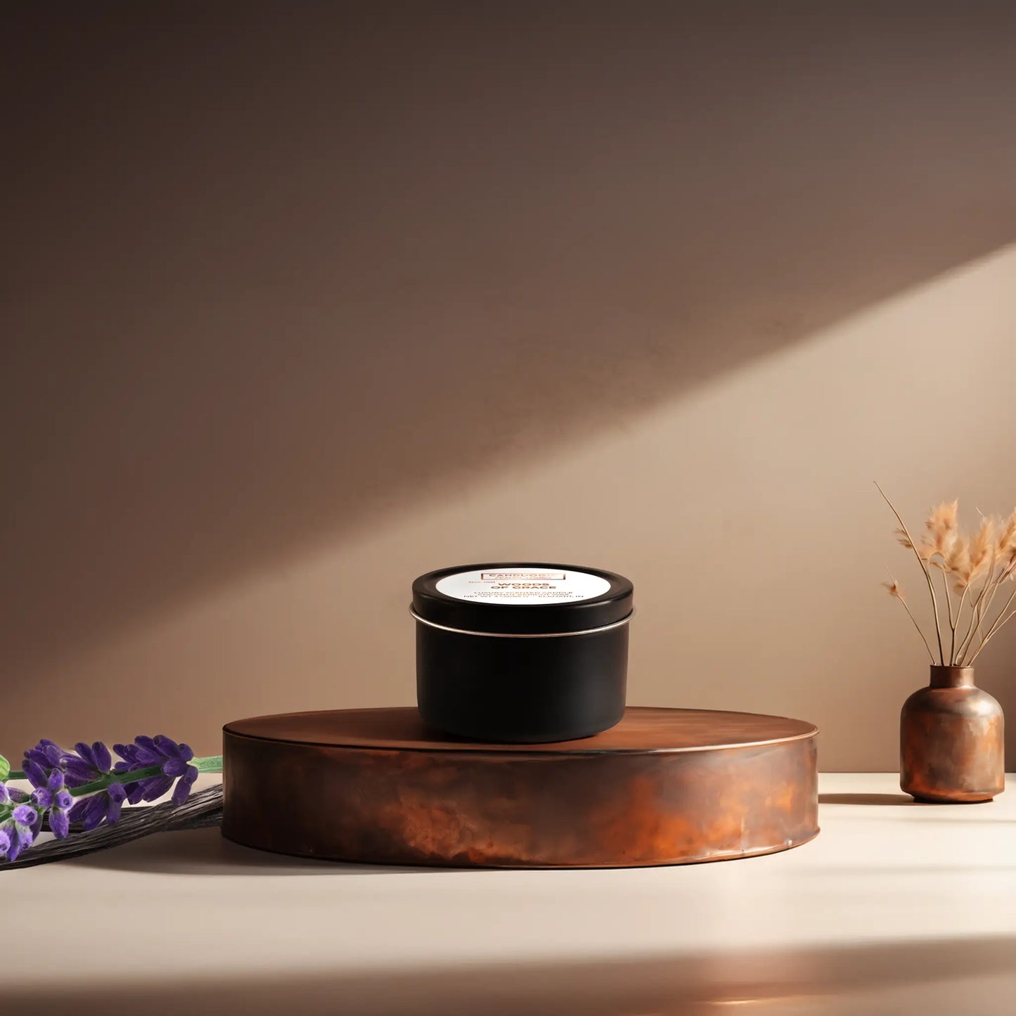 Candl•ogic’s scented candle with a wood wick in a 3 oz. black metal tin labeled ‘No. 001 Woods of Grace’ on a copper tray with lavender and vanilla beans and a copper vase in the background.