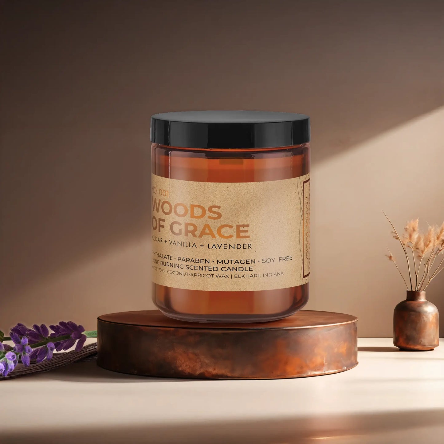 Candl•ogic’s scented candle with a wood wick in an amber glass 9oz. jar labeled ‘No. 001 Woods of Grace’ on a copper tray with lavender and vanilla beans and a copper vase in the background.
