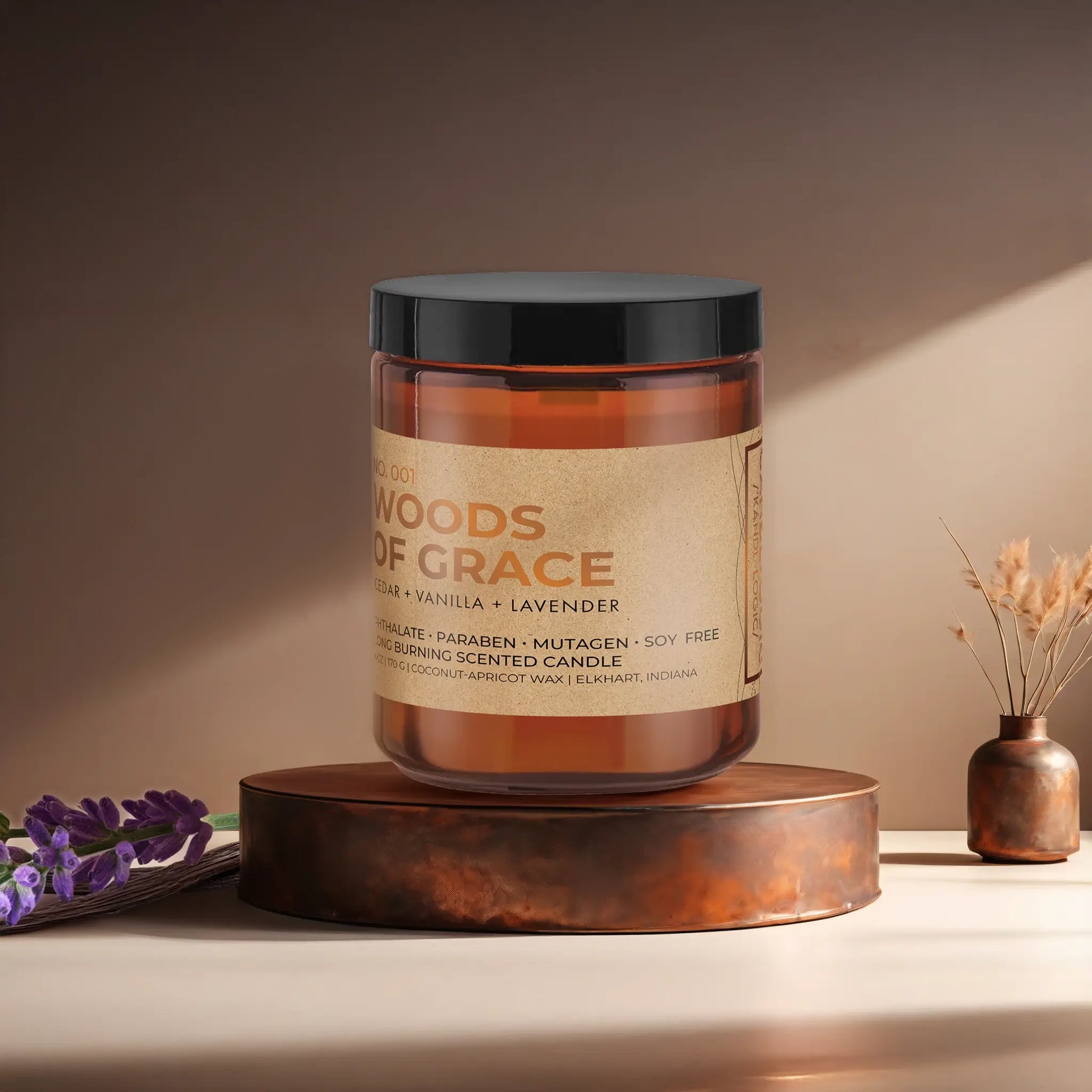 Candl•ogic’s scented candle with a wood wick in an amber glass 9oz. jar labeled ‘No. 001 Woods of Grace’ on a copper tray with lavender and vanilla beans and a copper vase in the background.
