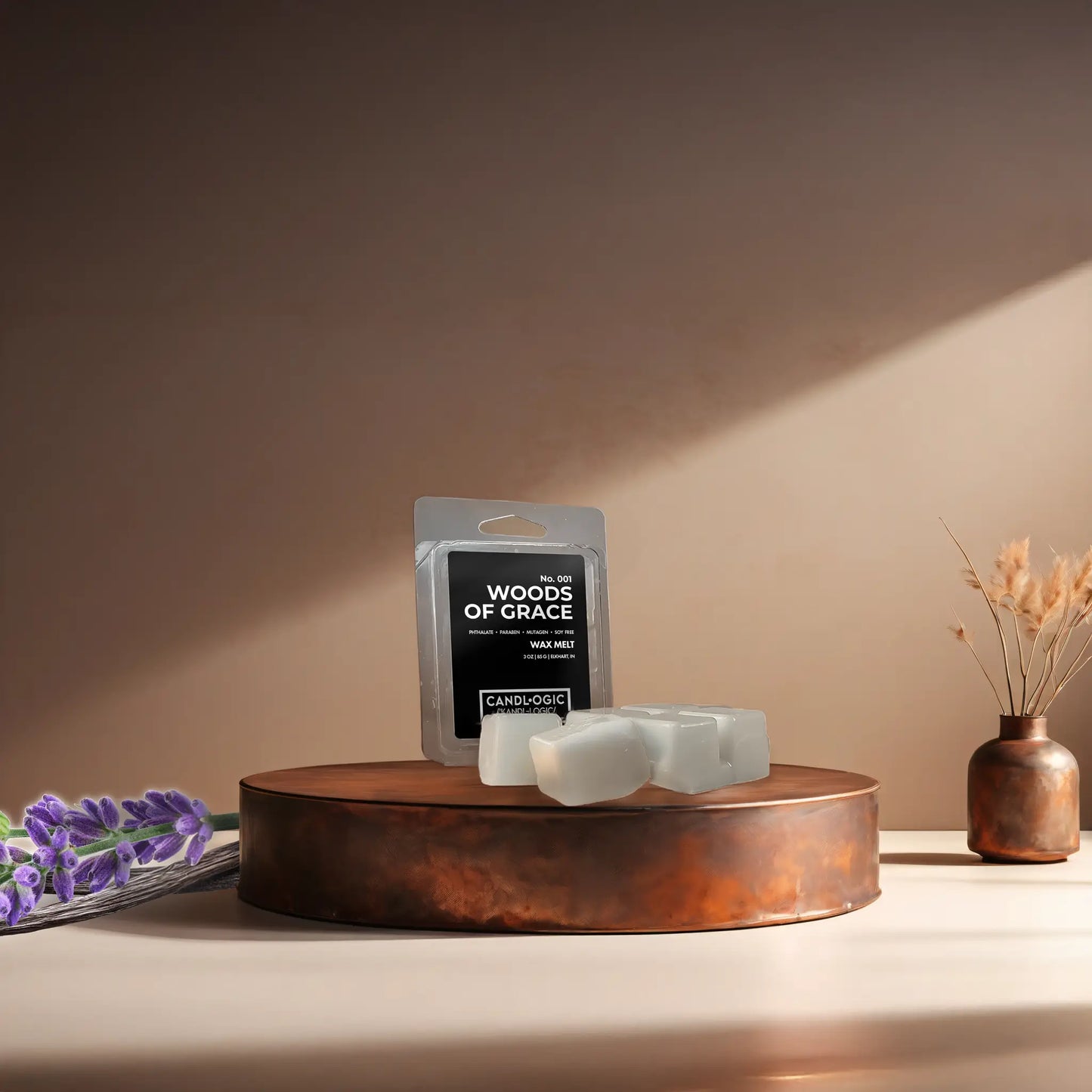 Candl•ogic’s wax melt cubes with packaging labeled ‘No. 001 Woods of Grace’ on a copper tray with lavender and vanilla beans and a copper vase in the background.
