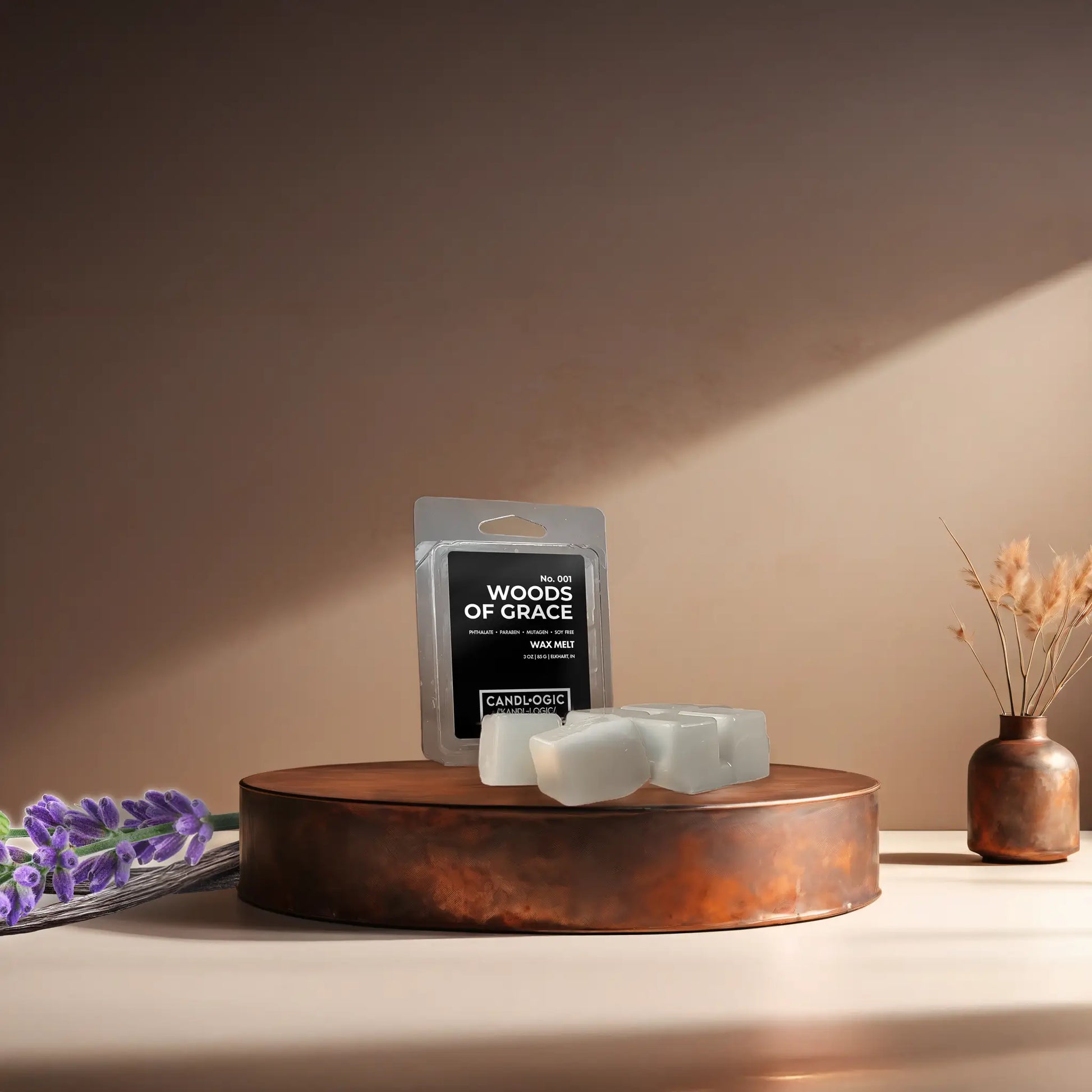 Candl•ogic’s wax melt cubes with packaging labeled ‘No. 001 Woods of Grace’ on a copper tray with lavender and vanilla beans and a copper vase in the background.
