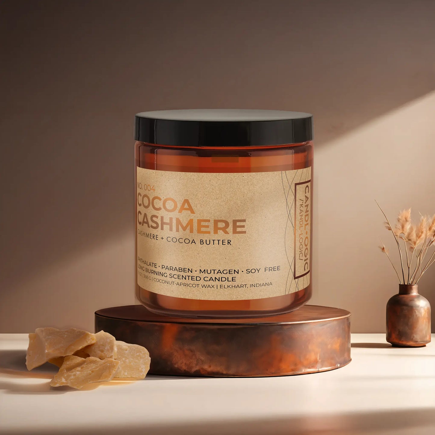 Candl•ogic’s scented candle with a wood wick in an amber glass 16 oz. jar labeled ‘No. 004 Cocoa Cashmere’ on a copper tray with an chunks of cocoa butter and a copper vase in the background.