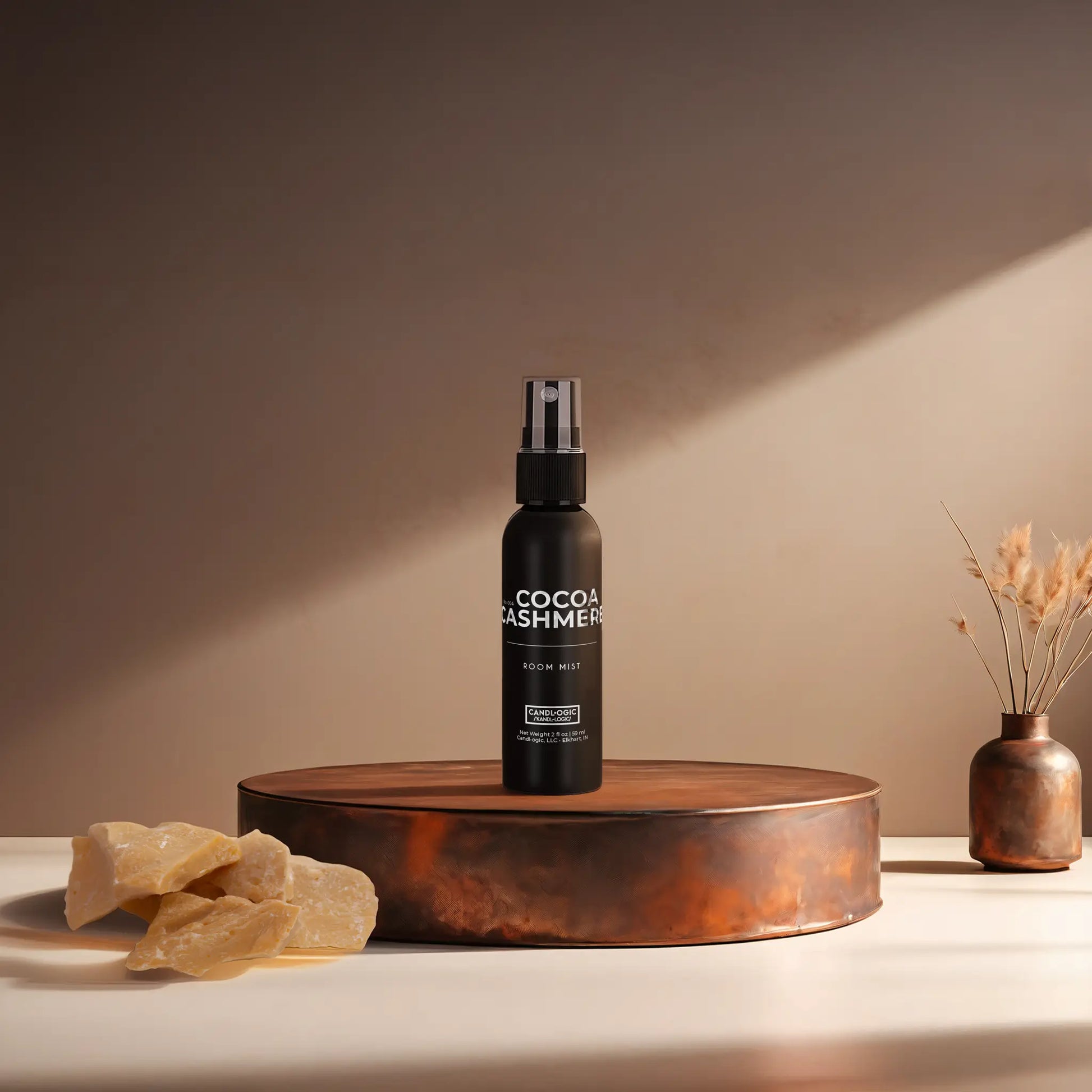 Candl•ogic’s 2 oz. room mist in a black spray bottle labeled ‘No. 004 Cocoa Cashmere’ on a copper tray with an chunks of cocoa butter and a copper vase in the background.