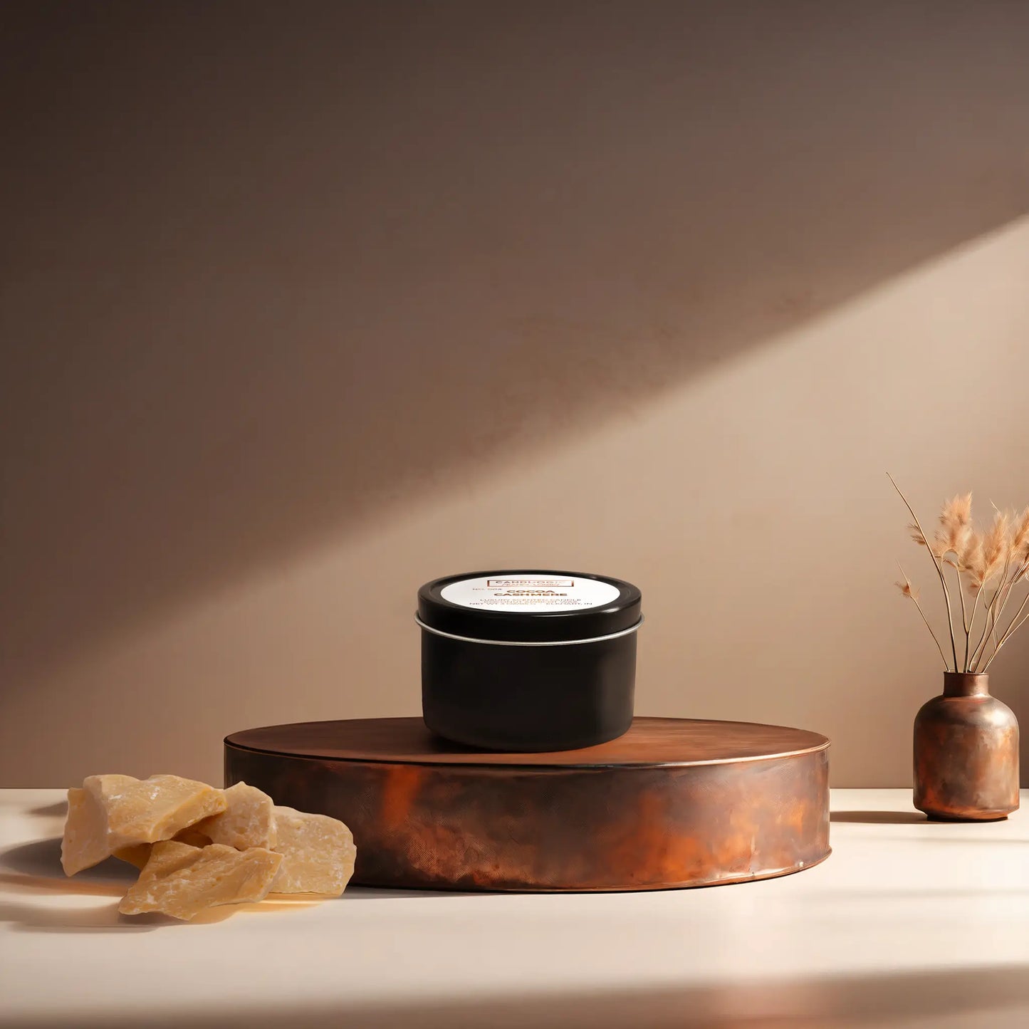 Candl•ogic’s scented candle with a wood wick in a 3 oz. black metal tin labeled ‘No. 004 Cocoa Cashmere’ on a copper tray with an chunks of cocoa butter and a copper vase in the background.