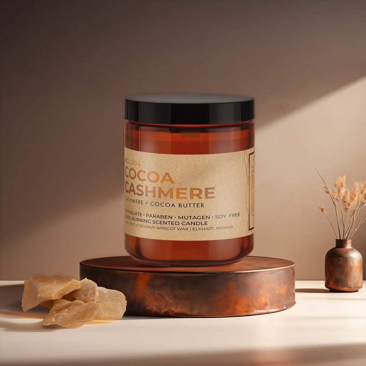 Candl•ogic’s scented candle with a wood wick in an amber glass 9 oz. jar labeled ‘No. 004 Cocoa Cashmere’ on a copper tray with an chunks of cocoa butter and a copper vase in the background.