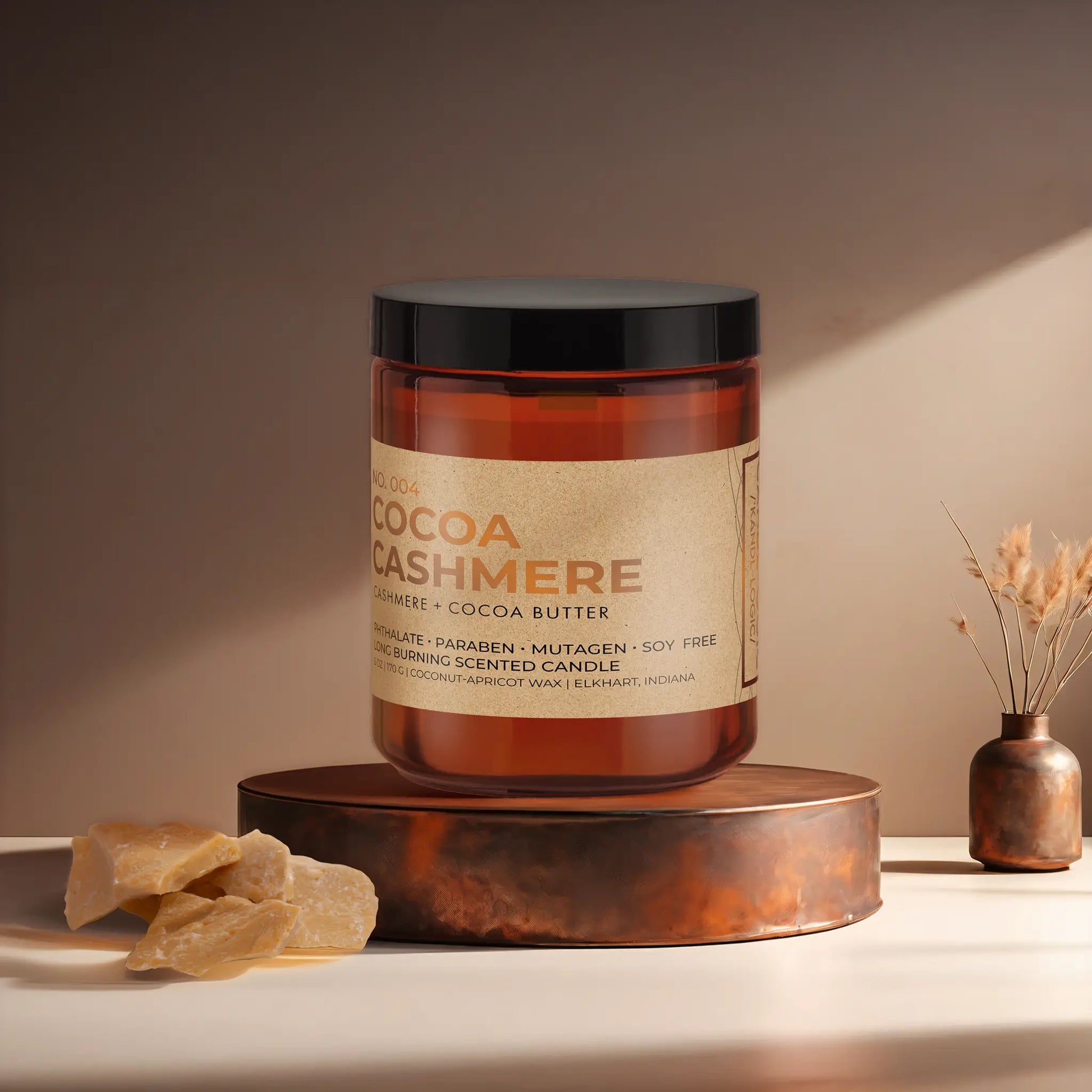 Candl•ogic’s scented candle with a wood wick in an amber glass 9 oz. jar labeled ‘No. 004 Cocoa Cashmere’ on a copper tray with an chunks of cocoa butter and a copper vase in the background.