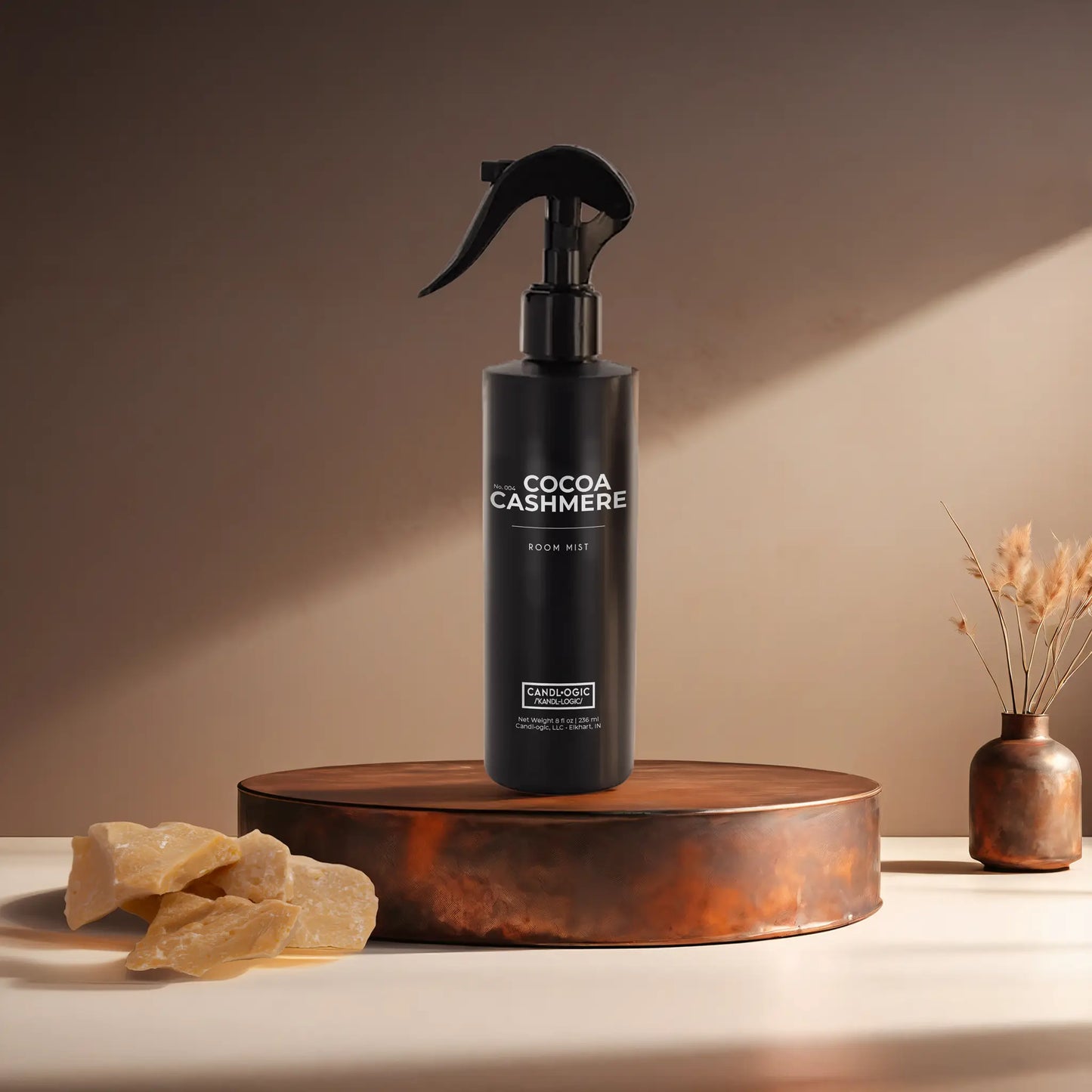 Candl•ogic’s 8 oz. room mist in a black spray bottle labeled ‘No. 004 Cocoa Cashmere’ on a copper tray with an chunks of cocoa butter and a copper vase in the background.