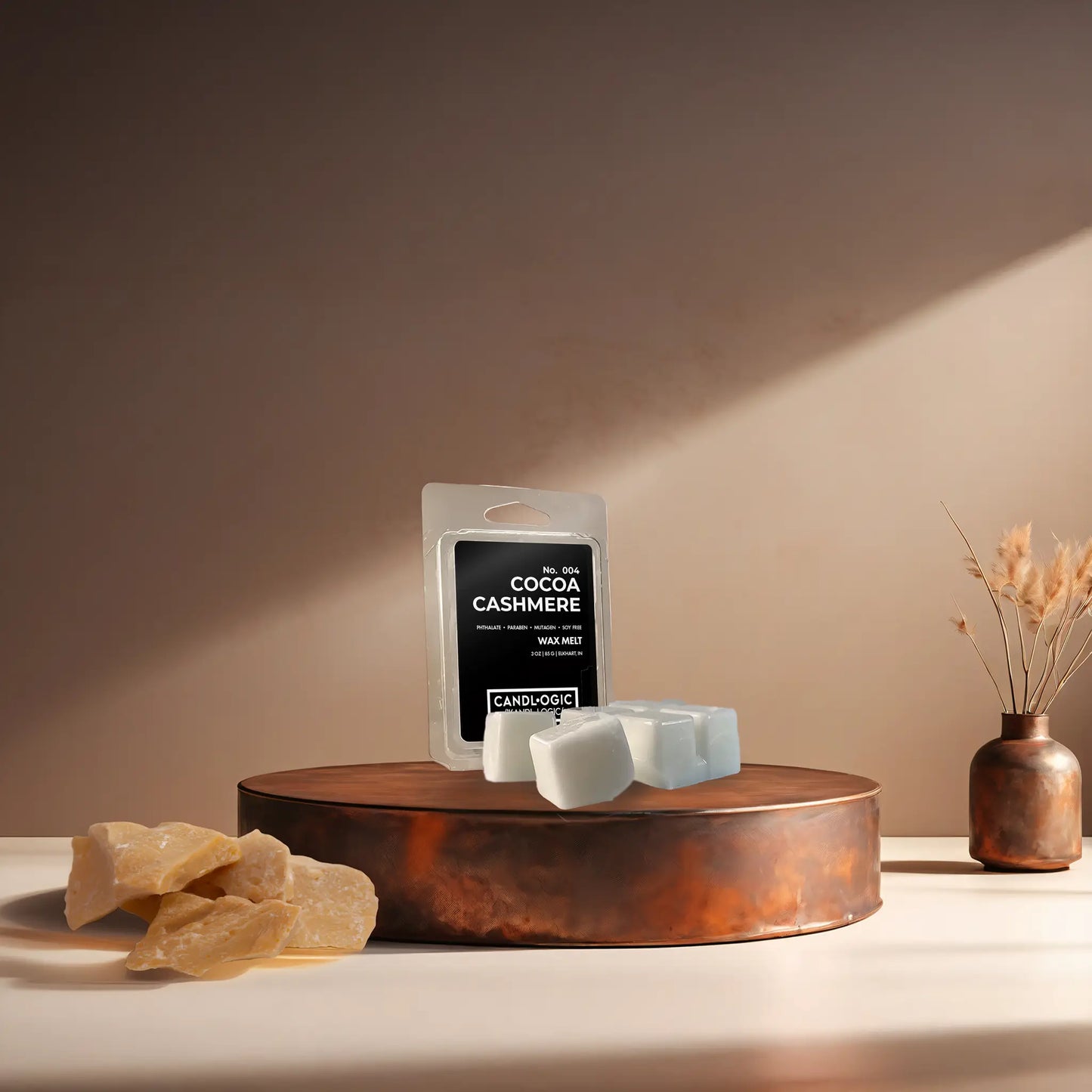 Candl•ogic’s wax melt cubes with packaging labeled ‘No. 004 Cocoa Cashmere’ on a copper tray with an chunks of cocoa butter and a copper vase in the background.