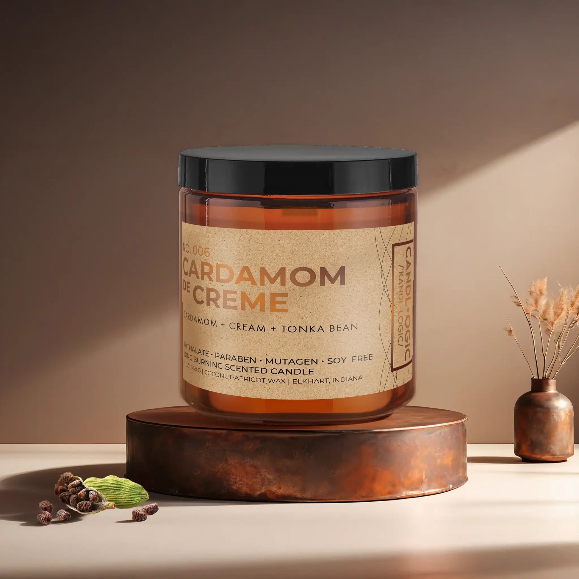 Candl•ogic’s scented candle with a wood wick in an amber glass 16 oz. jar labeled ‘No. 006 Cardamom de Creme’ on a copper tray with several black cardamom  and a copper vase in the background.