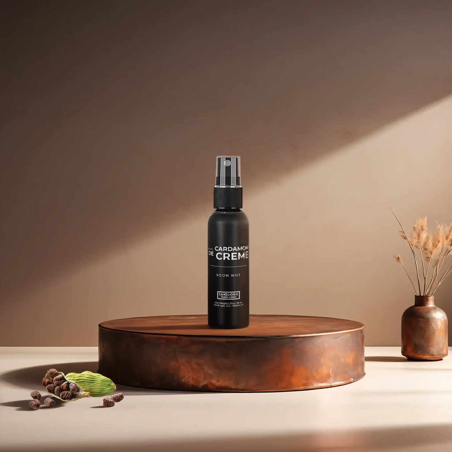Candl•ogic’s 2 oz. room mist in a black spray bottle labeled ‘No. 006 Cardamom de Creme’ on a copper tray with several black cardamom pods and a copper vase in the background.
