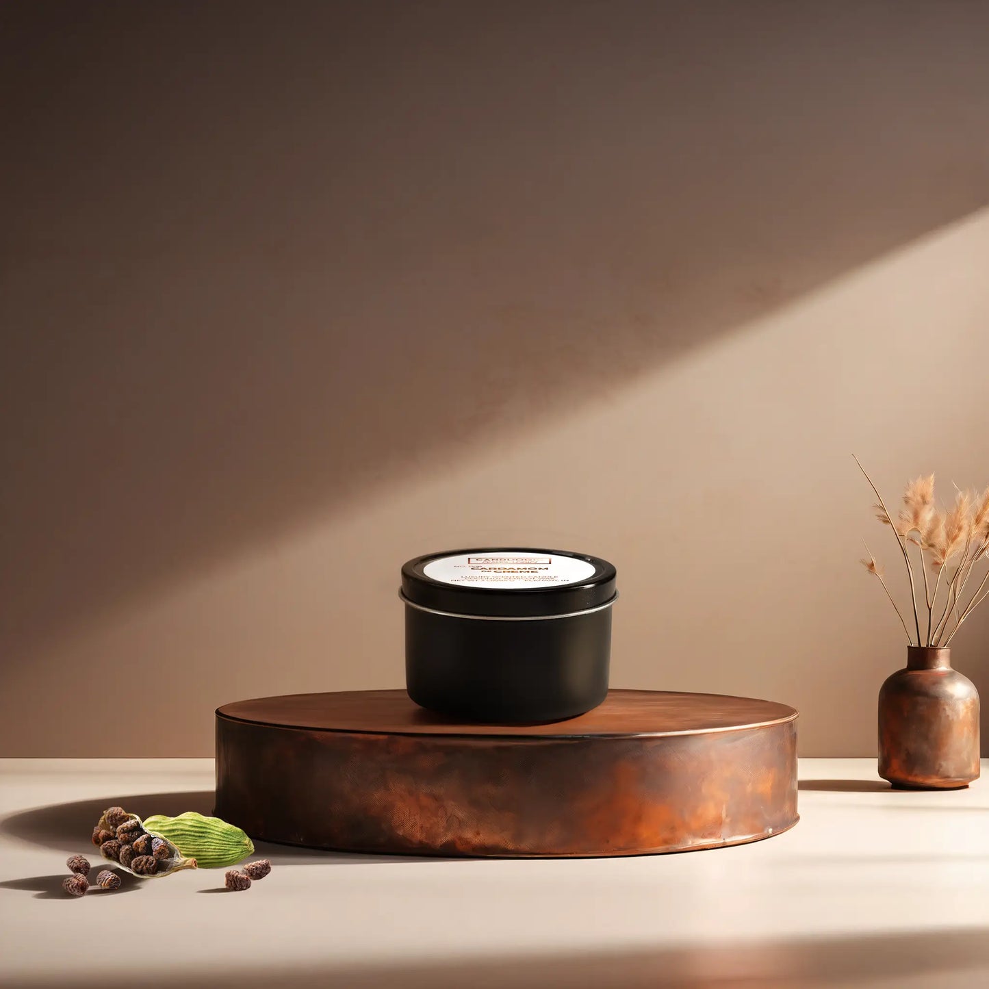 Candl•ogic’s scented candle with a wood wick in a 3 oz. black metla tin labeled ‘No. 006 Cardamom de Creme’ on a copper tray with several black cardamom  and a copper vase in the background.