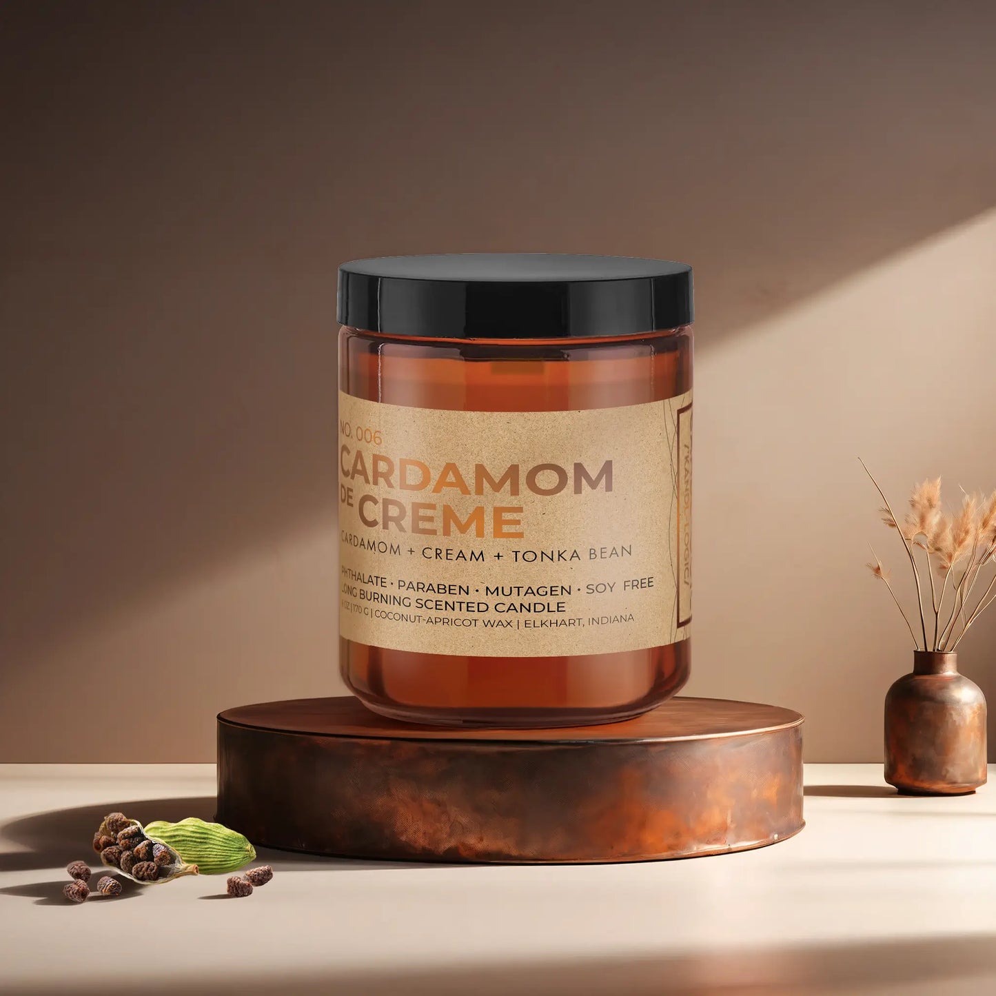 Candl•ogic’s scented candle with a wood wick in an amber glass 9 oz. jar labeled ‘No. 006 Cardamom de Creme’ on a copper tray with several black cardamom  and a copper vase in the background.
