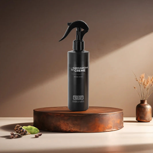 Candl•ogic’s 8 oz. room mist in a black spray bottle labeled ‘No. 006 Cardamom de Creme’ on a copper tray with several black cardamom pods and a copper vase in the background.