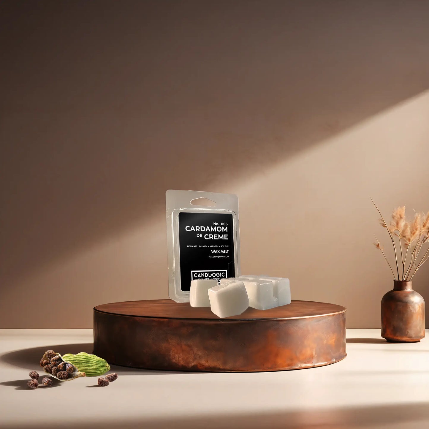 Candl•ogic’s wax melt cubes with packaging labeled ‘No. 006 Cardamom de Creme’ on a copper tray with several black cardamom pods and a copper vase in the background.