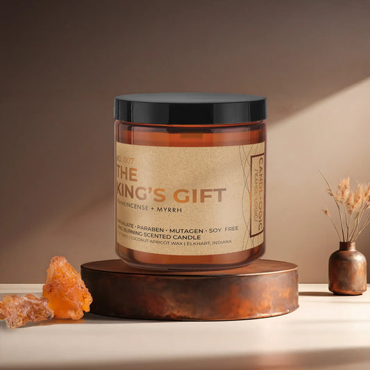 Candl•ogic’s scented candle with a wood wick in an amber glass 16 oz. jar labeled ‘No. 007 The King’s Gift’ on a copper tray with frankincense and myrrh and a copper vase in the background.
