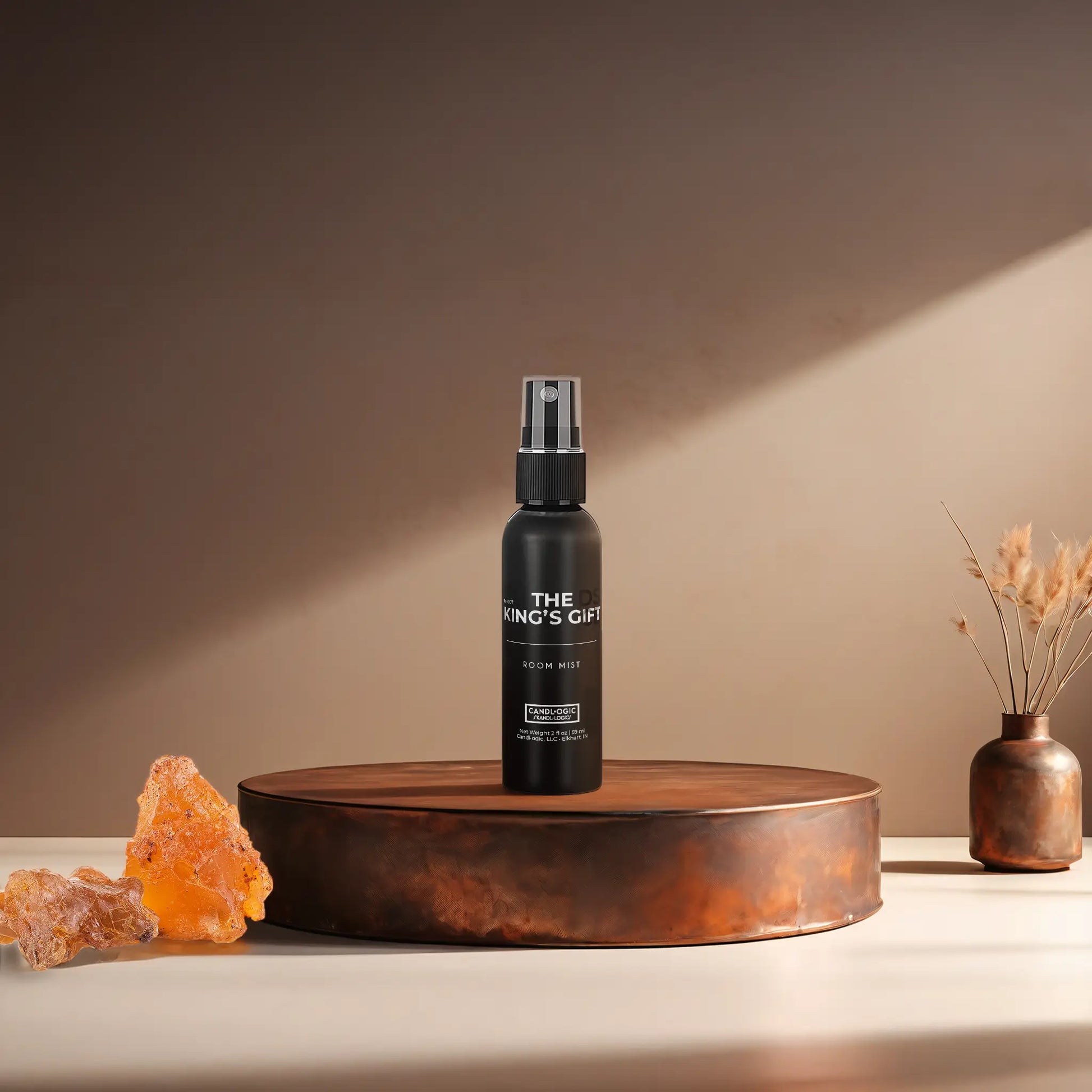 Candl•ogic’s 2 oz. room mist in a black spray bottle labeled ‘No. 007 The King’s Gift’ on a copper tray with frankincense and myrrh and a copper vase in the background.