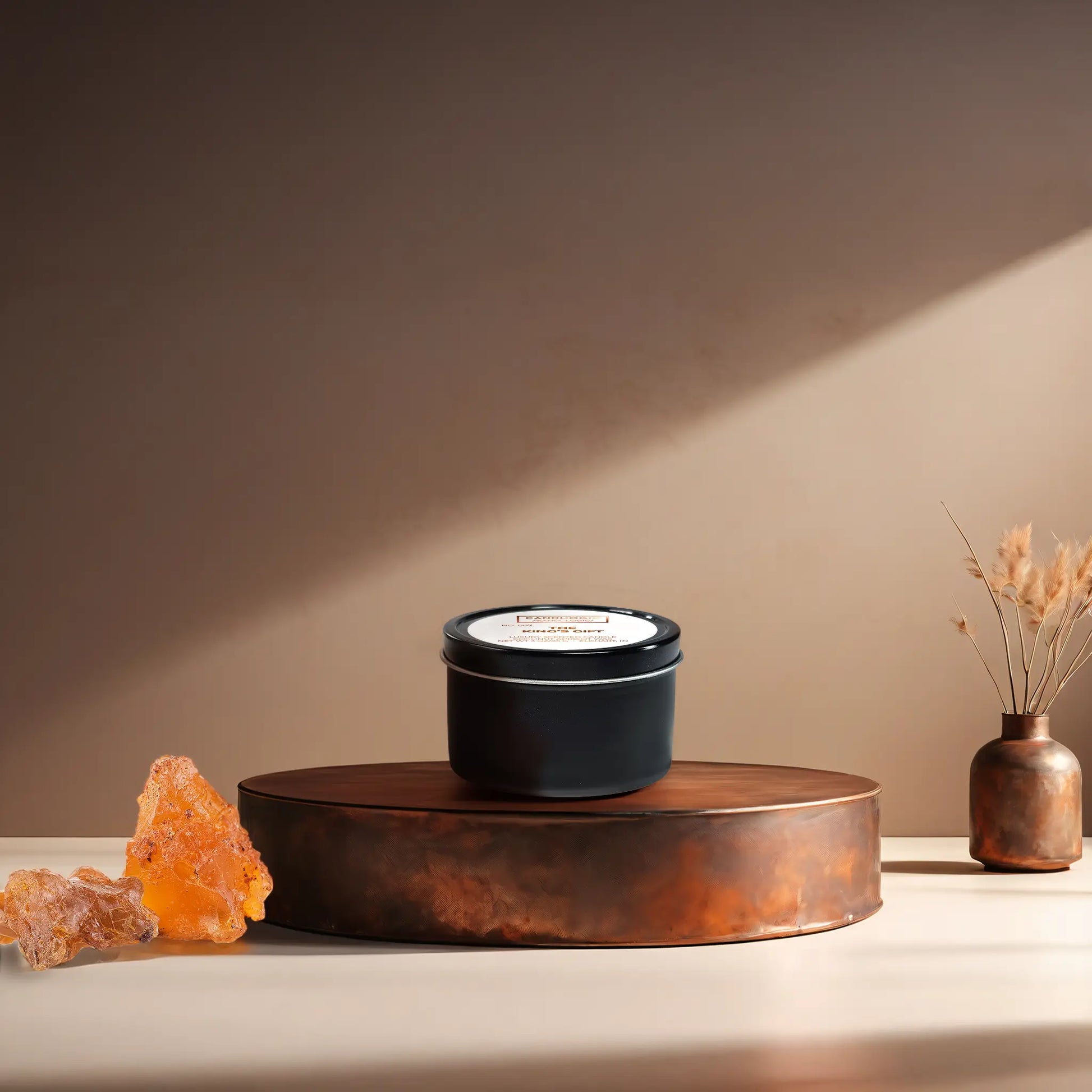 Candl•ogic’s scented candle with a wood wick in a 3 oz. black metla tin labeled ‘No. 007 The King’s Gift’ on a copper tray with frankincense and myrrh and a copper vase in the background.