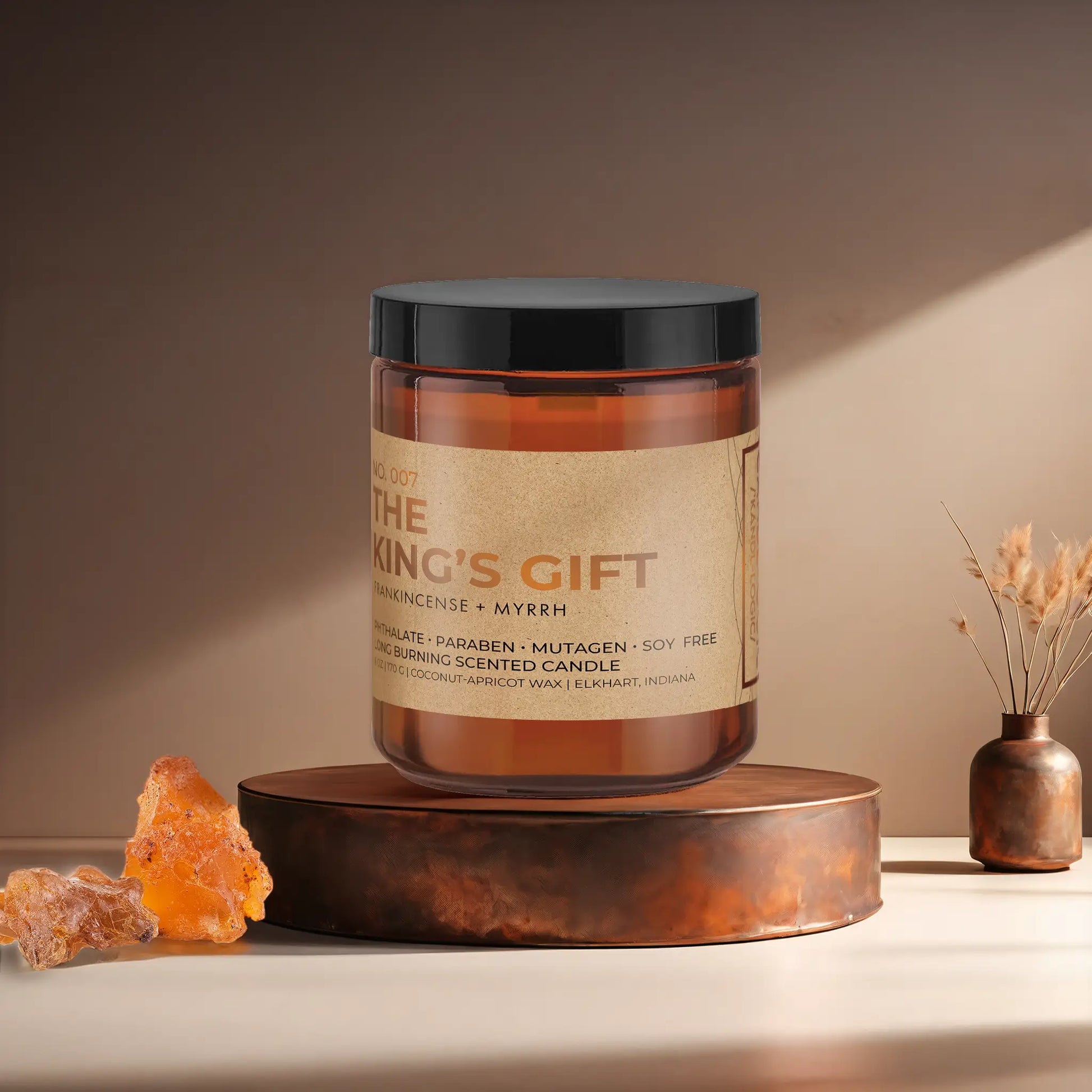 Candl•ogic’s scented candle with a wood wick in an amber glass 9 oz. jar labeled ‘No. 007 The King’s Gift’ on a copper tray with frankincense and myrrh and a copper vase in the background.