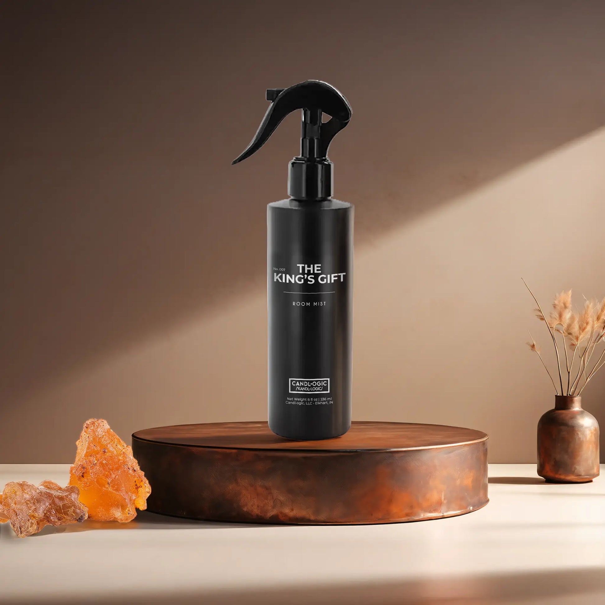 Candl•ogic’s 8 oz. room mist in a black spray bottle labeled ‘No. 007 The King’s Gift’ on a copper tray with frankincense and myrrh and a copper vase in the background.