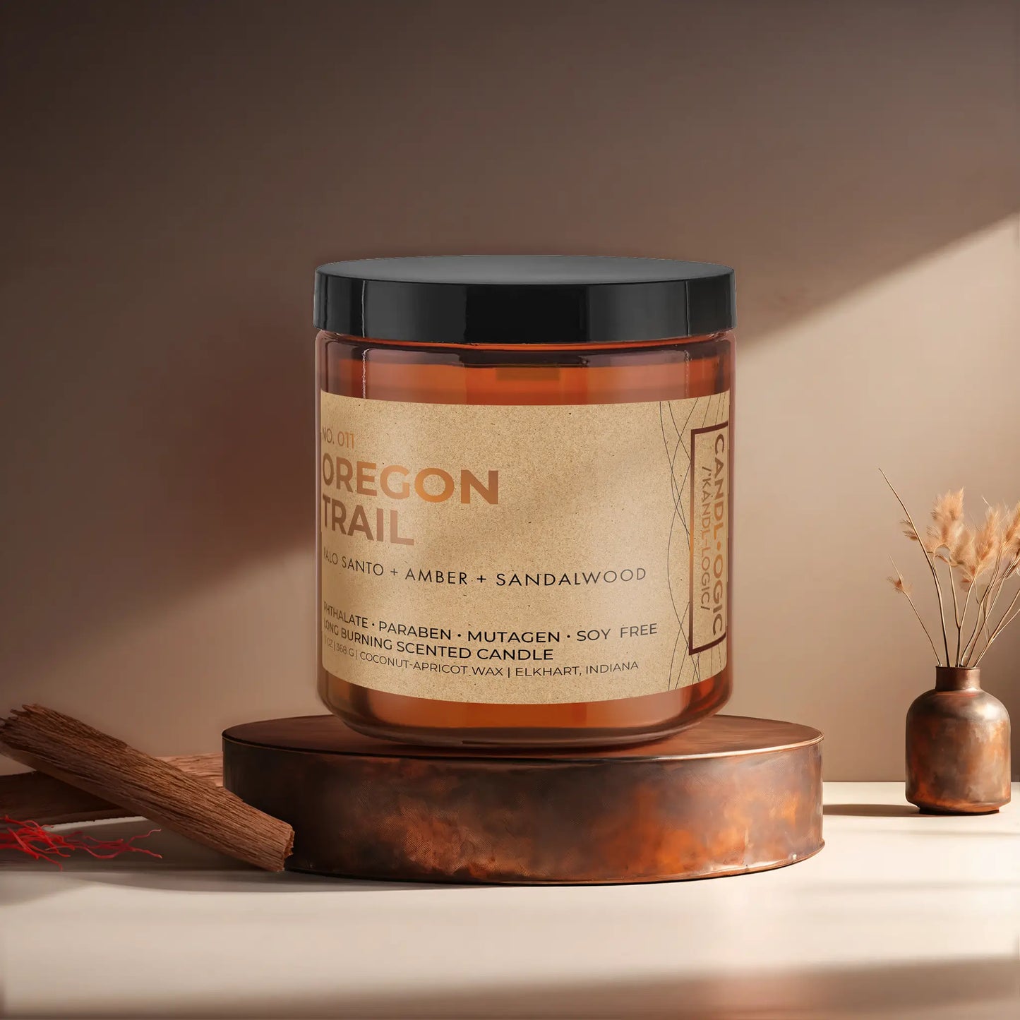 Candl•ogic’s scented candle with a wood wick in an amber glass 16 oz. jar labeled ‘No. 011 Oregon Trail’ on a copper tray with pieces of sandalwood and strands of saffon and a copper vase in the background.
