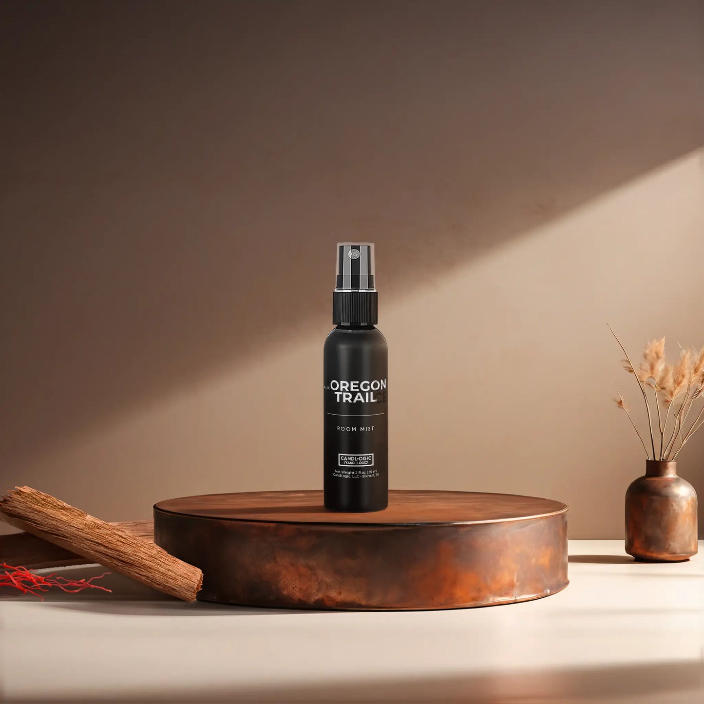 Candl•ogic’s 2 oz. room mist in a black spray bottle labeled ‘No. 011 Oregon Trail’ on a copper tray with pieces of sandalwood and strands of saffon and a copper vase in the background.