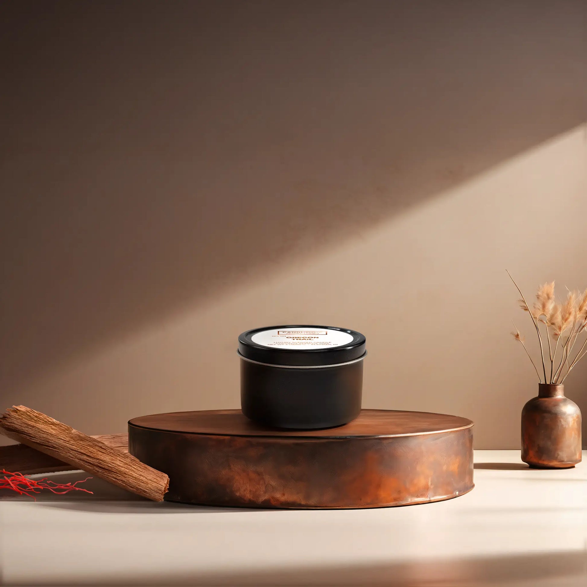 Candl•ogic’s scented candle with a wood wick in a 3 oz. black metal tin labeled ‘No. 011 Oregon Trail’ on a copper tray with pieces of sandalwood and strands of saffon and a copper vase in the background.