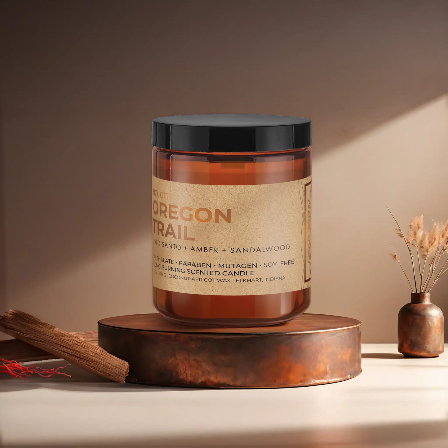 Candl•ogic’s scented candle with a wood wick in an amber glass 9 oz. jar labeled ‘No. 011 Oregon Trail’ on a copper tray with pieces of sandalwood and strands of saffon and a copper vase in the background.