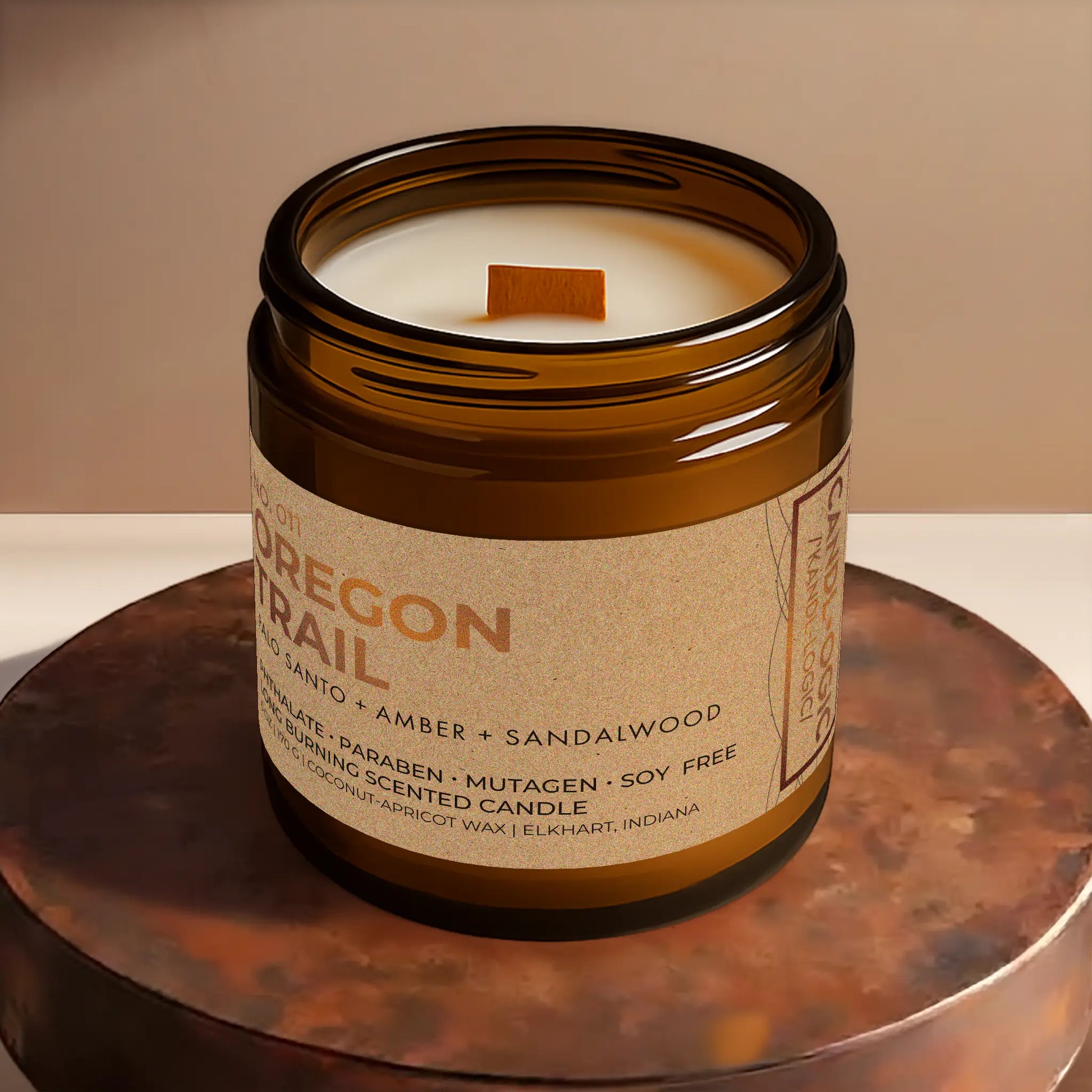 Candl•ogic’s scented candle with a wood wick viewed from the top in an amber glass 16 oz. jar labeled ‘No. 011 Oregon Trail’ on a copper tray.
