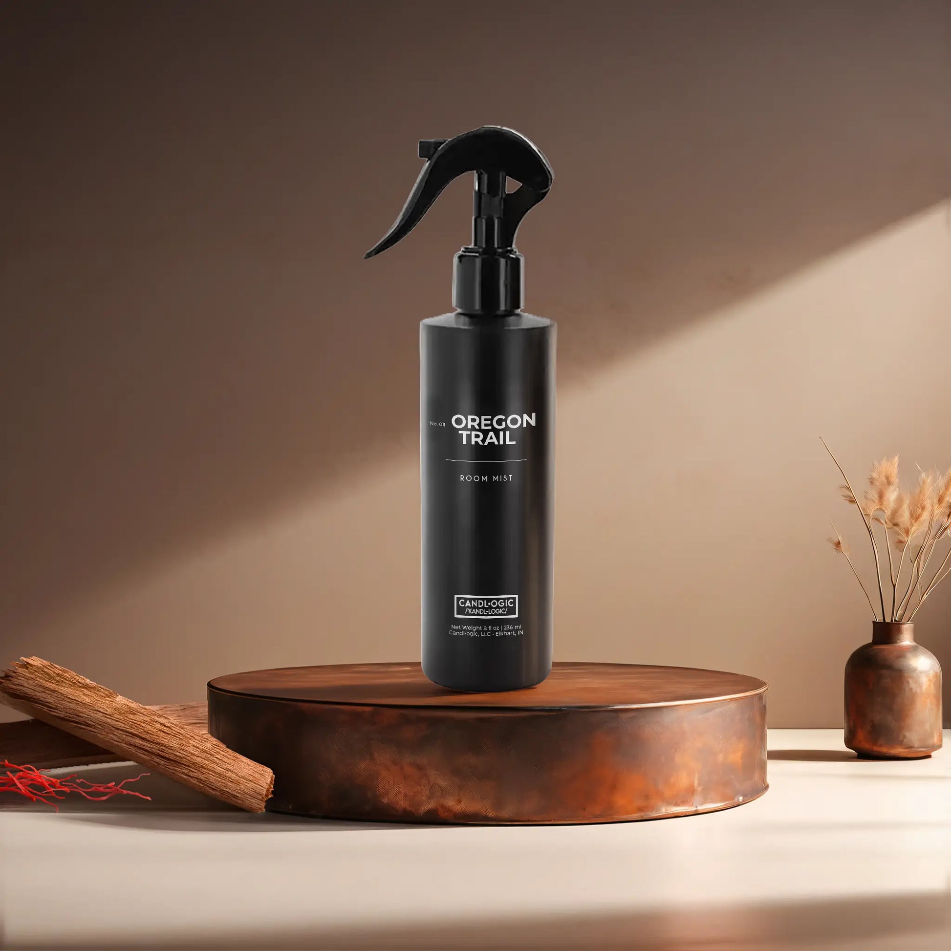 Candl•ogic’s 8 oz. room mist in a black spray bottle labeled ‘No. 011 Oregon Trail’ on a copper tray with pieces of sandalwood and strands of saffon and a copper vase in the background.