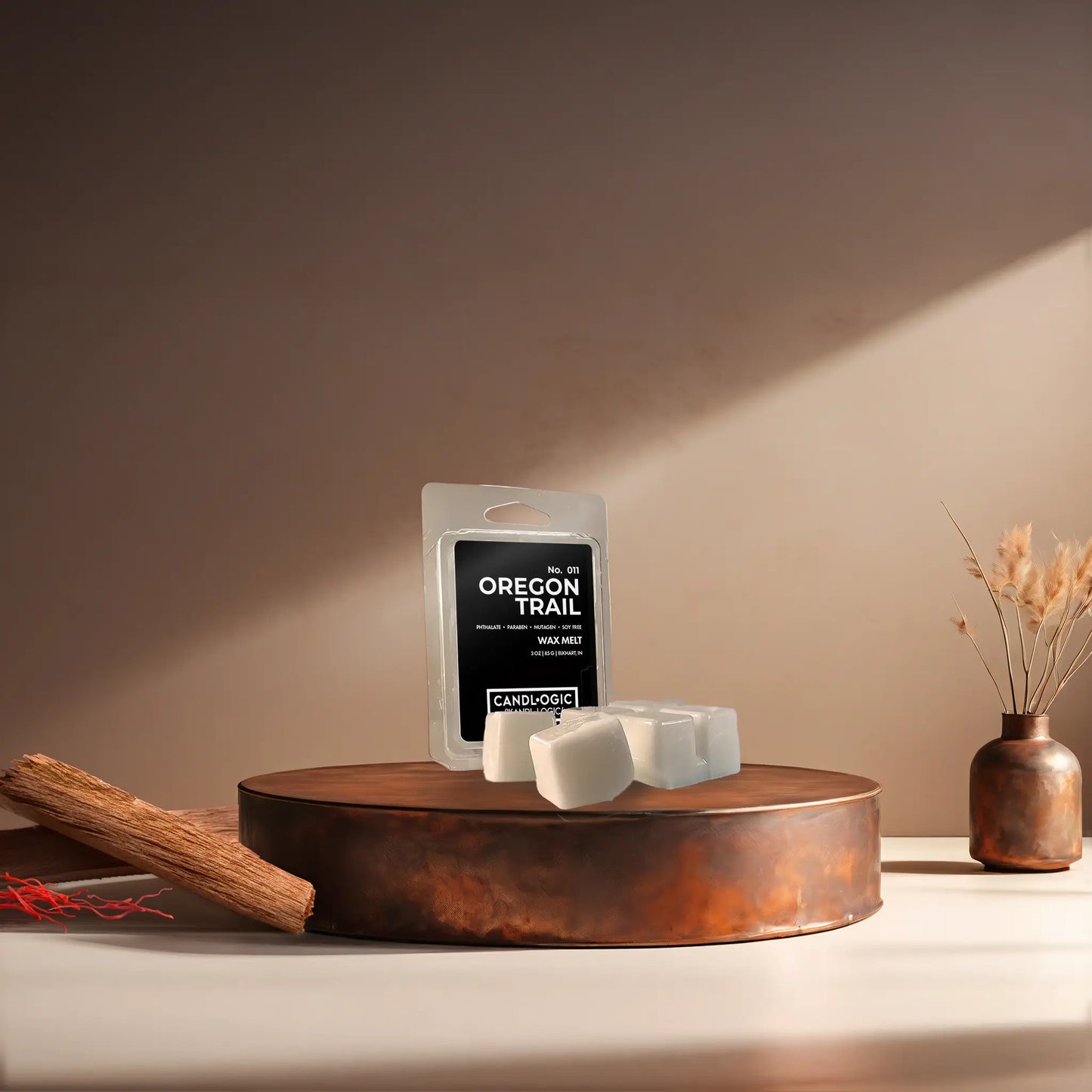 Candl•ogic’s wax melt cubes with packaging labeled ‘No. 011 Oregon Trail’ on a copper tray with pieces of sandalwood and strands of saffon and a copper vase in the background.