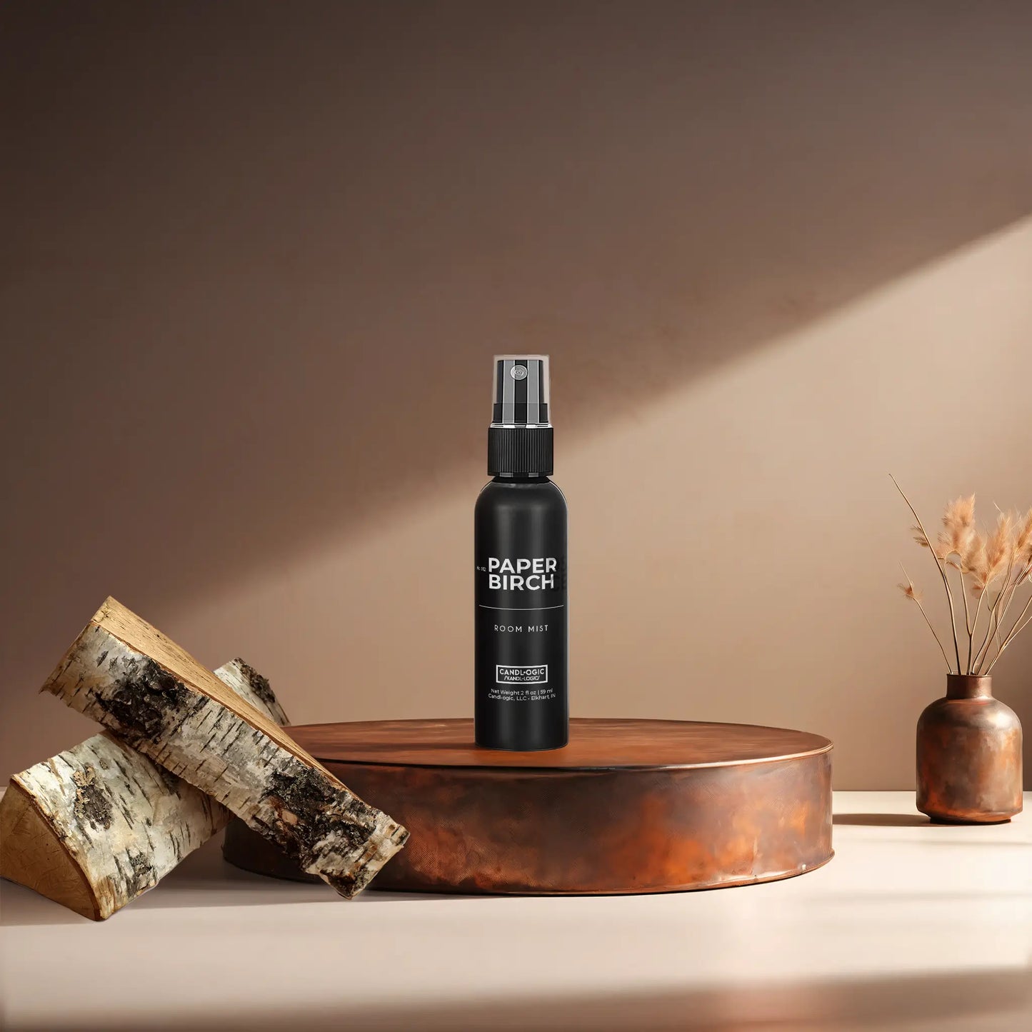 Candl•ogic’s 2 oz. room mist in a black spray bottle labeled ‘No. 012 Paper Birch’ on a copper tray with pieces of paper birch tree and a copper vase in the background.