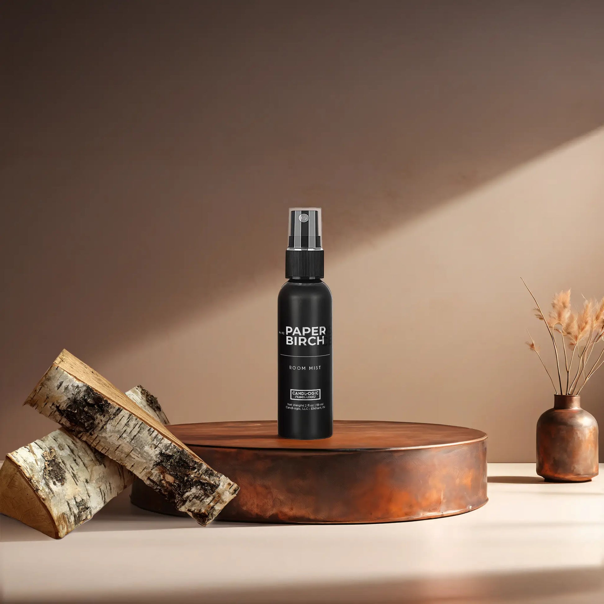 Candl•ogic’s 2 oz. room mist in a black spray bottle labeled ‘No. 012 Paper Birch’ on a copper tray with pieces of paper birch tree and a copper vase in the background.