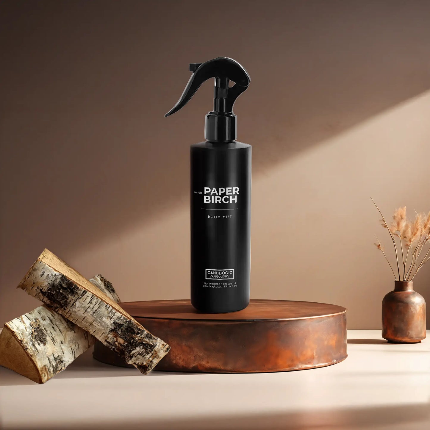 Candl•ogic’s 8 oz. room mist in a black spray bottle labeled ‘No. 012 Paper Birch’ on a copper tray with pieces of paper birch tree and a copper vase in the background.