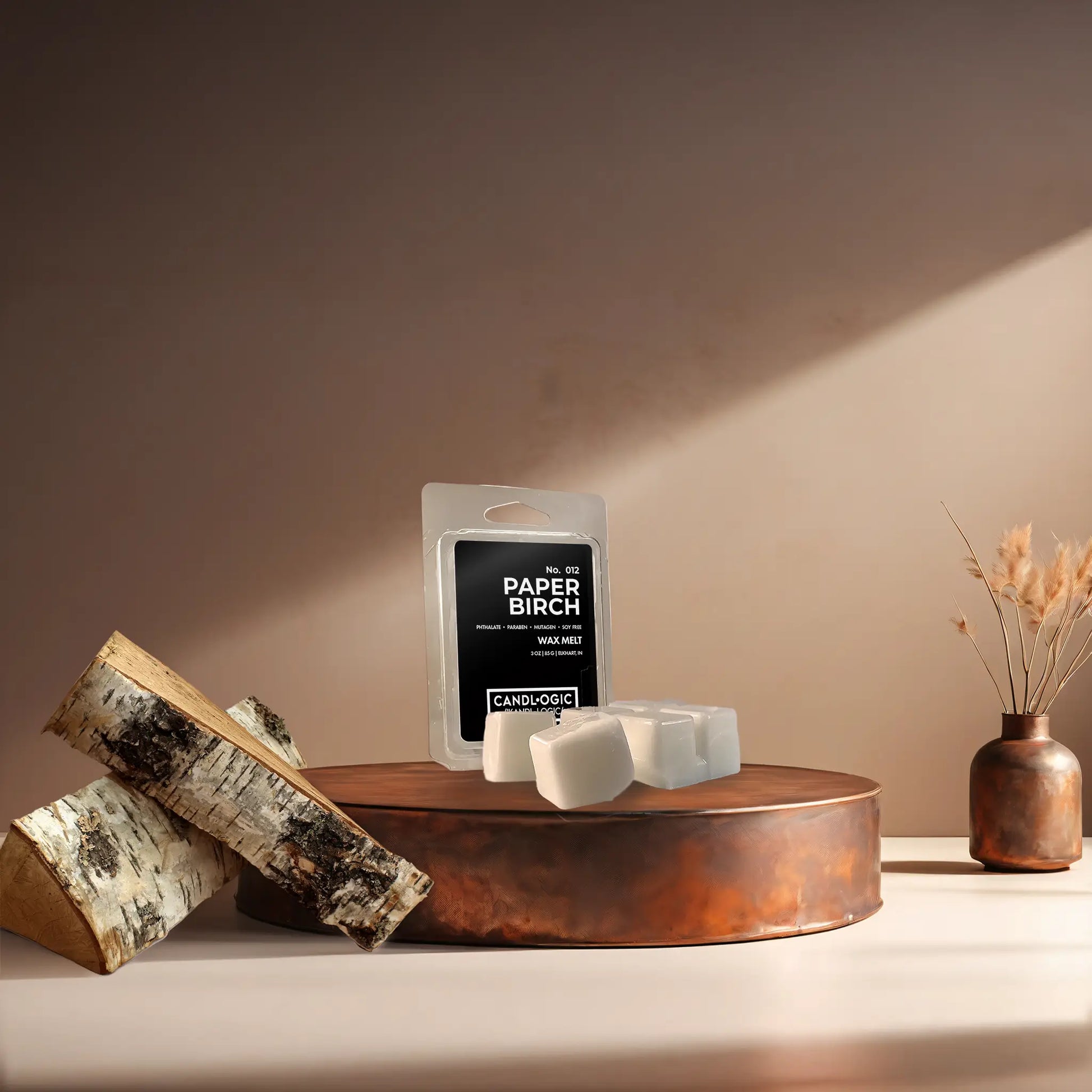 Candl•ogic’s wax melt cubes with packaging labeled ‘No. 012 Paper Birch’ on a copper tray with pieces of paper birch tree and a copper vase in the background.