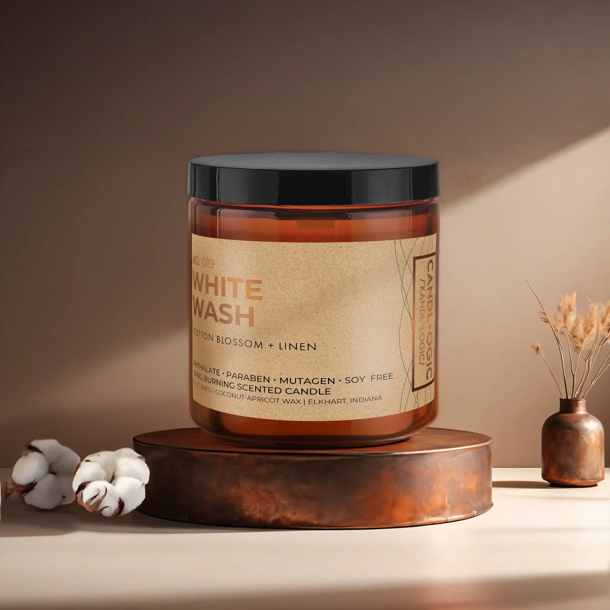 Candl•ogic’s scented candle with a wood wick in an amber glass 16 oz. jar labeled ‘No. 019 White Wash’ on a copper tray with cotton flower stems and a copper vase in the background.