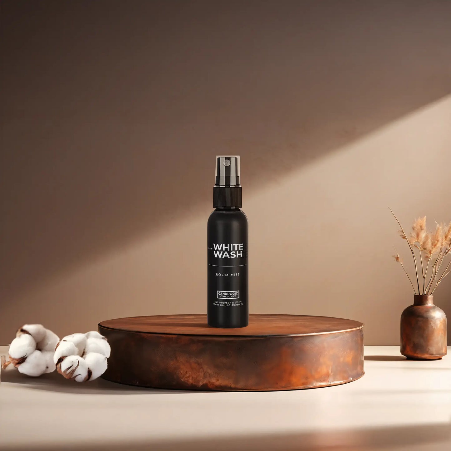 Candl•ogic’s 2 oz. room mist in a black spray bottle labeled ‘No. 019 White Wash’ on a copper tray with cotton flower stems and a copper vase in the background.