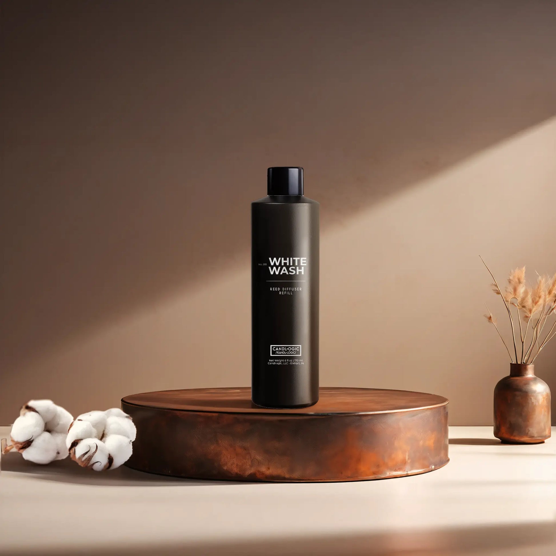 Candl•ogic’s 6 oz. reed diffuser refill solution in a black plastic bottle labeled ‘No. 019 White Wash’ on a copper tray with cotton flower stems and a copper vase in the background.