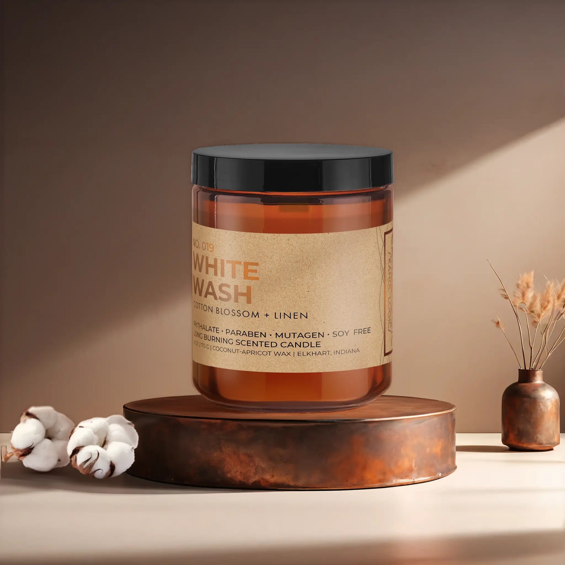Candl•ogic’s scented candle with a wood wick in an amber glass 9 oz. jar labeled ‘No. 019 White Wash’ on a copper tray with cotton flower stems and a copper vase in the background.