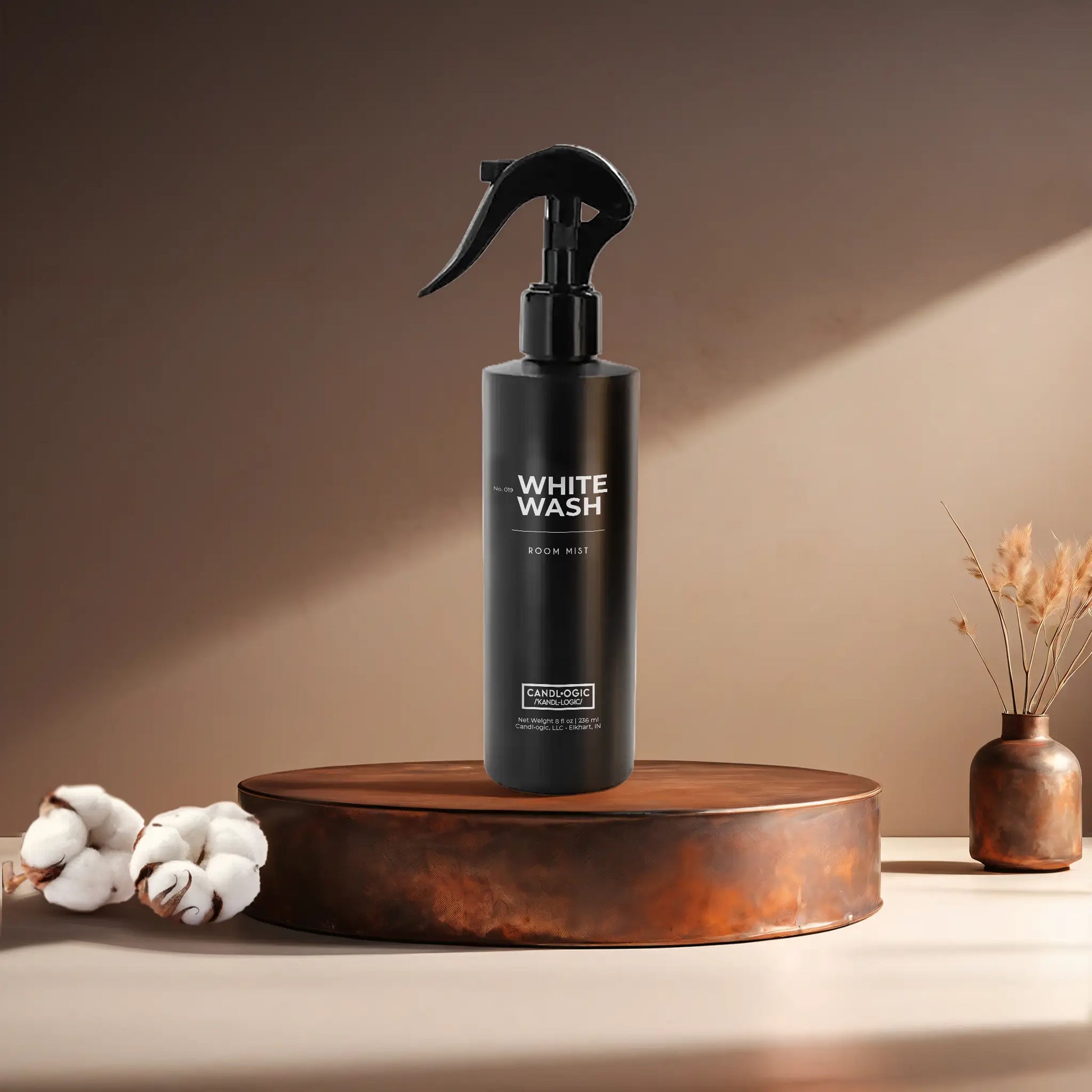 Candl•ogic’s 8 oz. room mist in a black spray bottle labeled ‘No. 019 White Wash’ on a copper tray with cotton flower stems and a copper vase in the background.