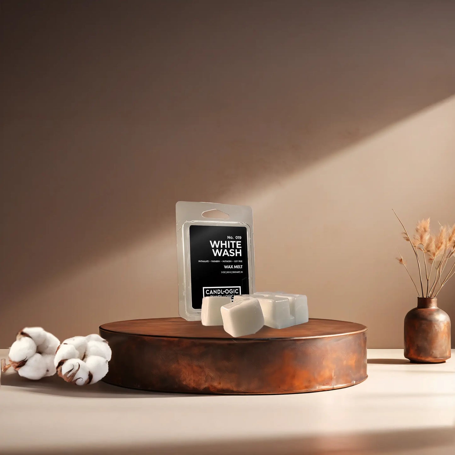 Candl•ogic’s wax melt cubes with packaging labeled ‘No. 019 White Wash’ on a copper tray with cotton flower stems and a copper vase in the background.