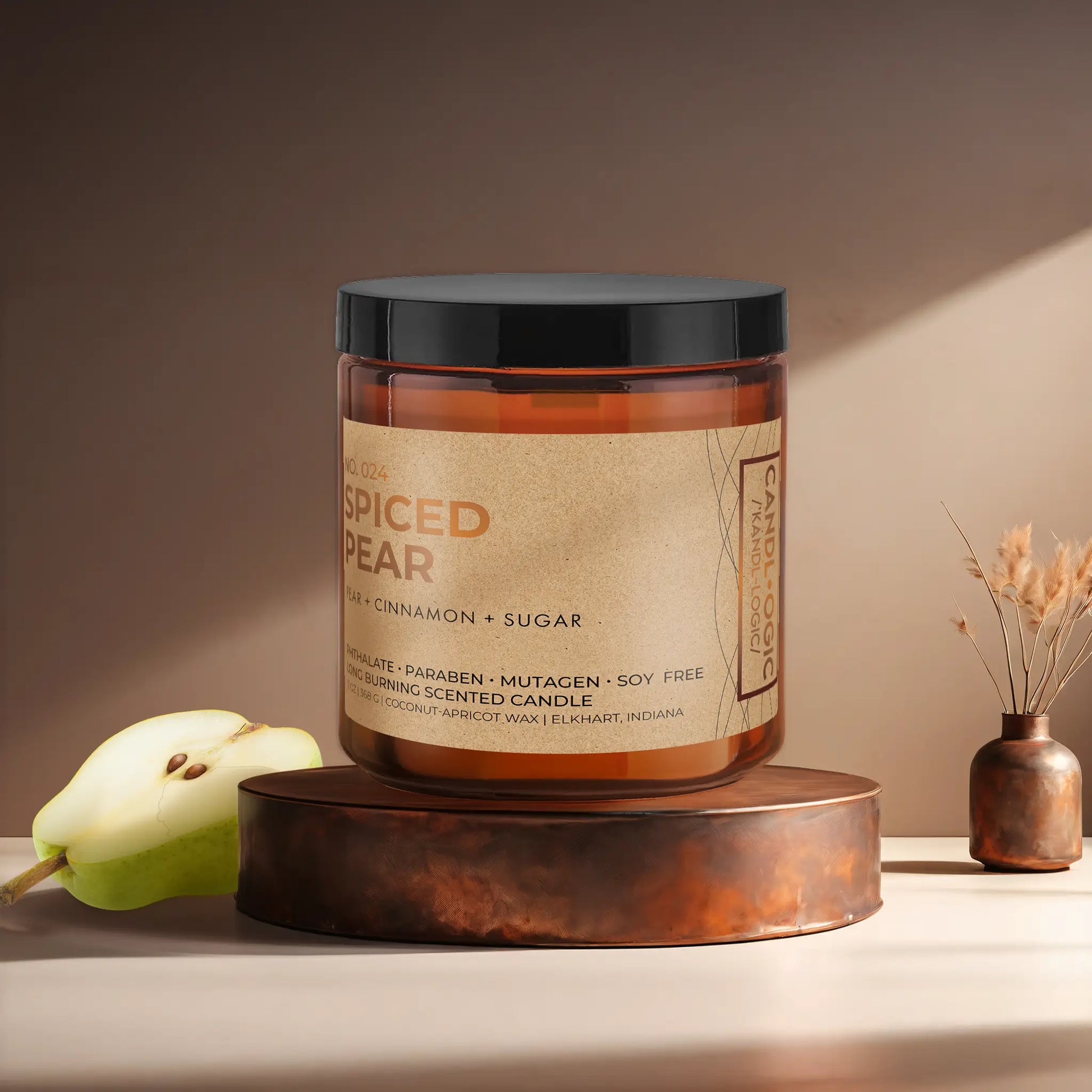 Candl•ogic’s scented candle with a wood wick in an amber glass 16 oz. jar labeled ‘No. 024 Spiced Pear’ on a copper tray with a sliced pear and a copper vase in the background.