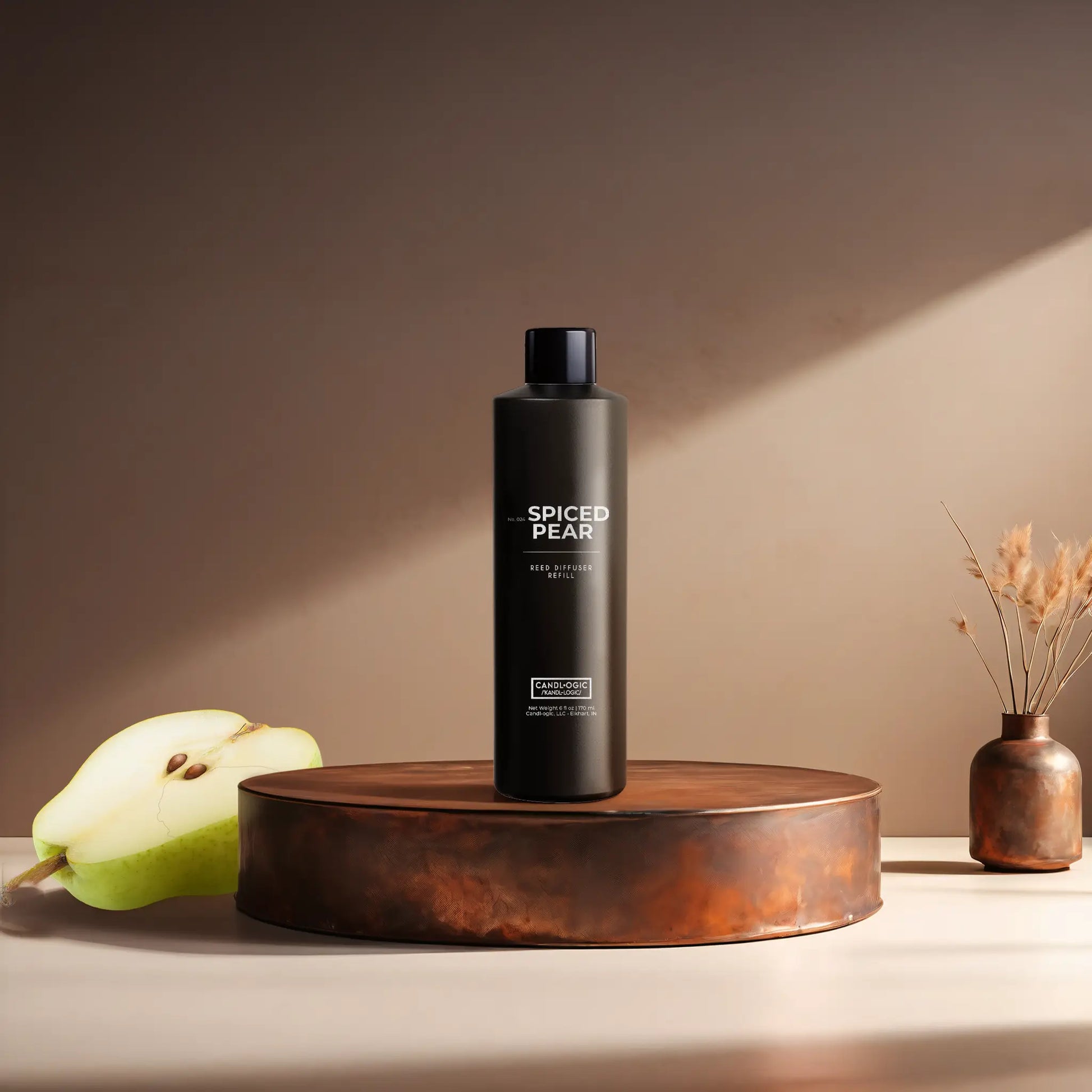 Candl•ogic’s 6 oz. reed diffuser refill solution in a black plastic bottle labeled ‘No. 024 Spiced Pear’ on a copper tray with a sliced pear and a copper vase in the background.