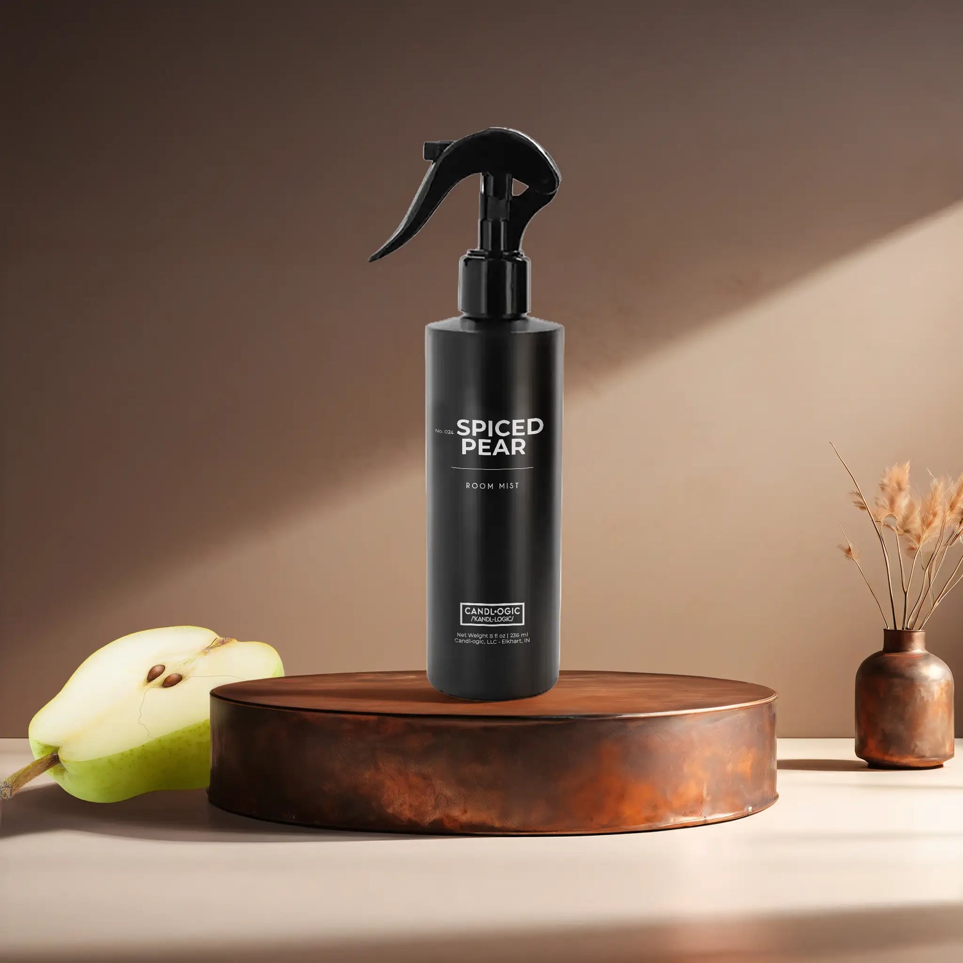Candl•ogic’s 8 oz. room mist in a black spray bottle labeled ‘No. 024 Spiced Pear’ on a copper tray with a sliced pear and a copper vase in the background.