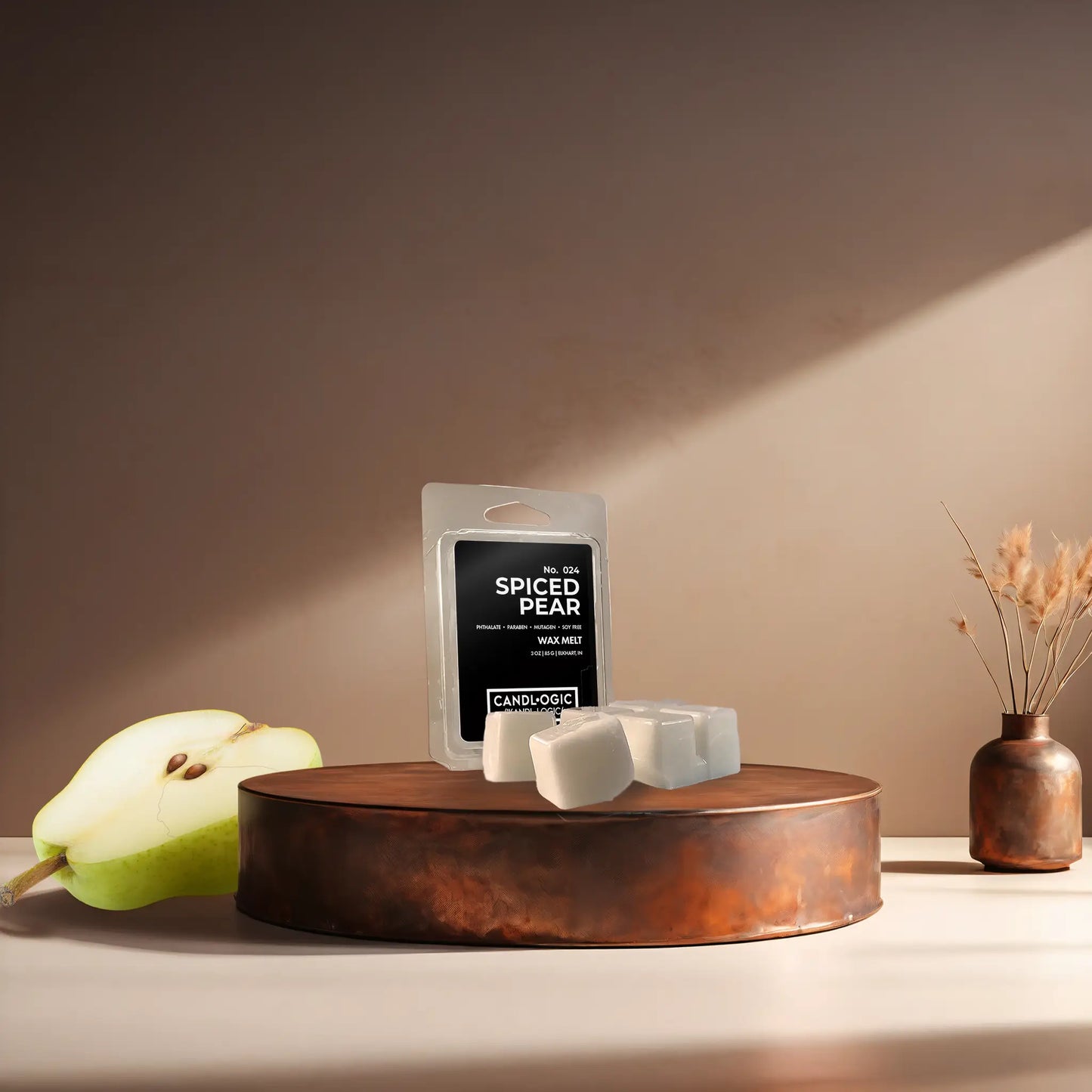 Candl•ogic’s wax melt cubes with packaging labeled ‘No. 024 Spiced Pear’ on a copper tray with a sliced pear and a copper vase in the background.
